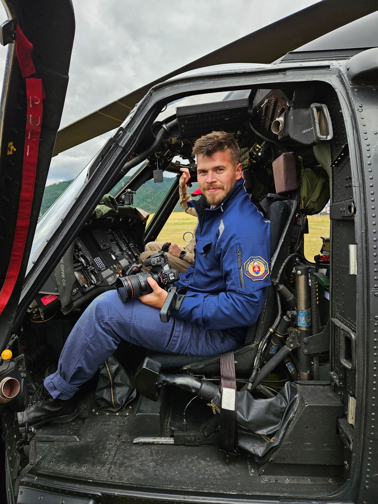 Pilot sits in helicopter cockpit wearing blue flight suit.