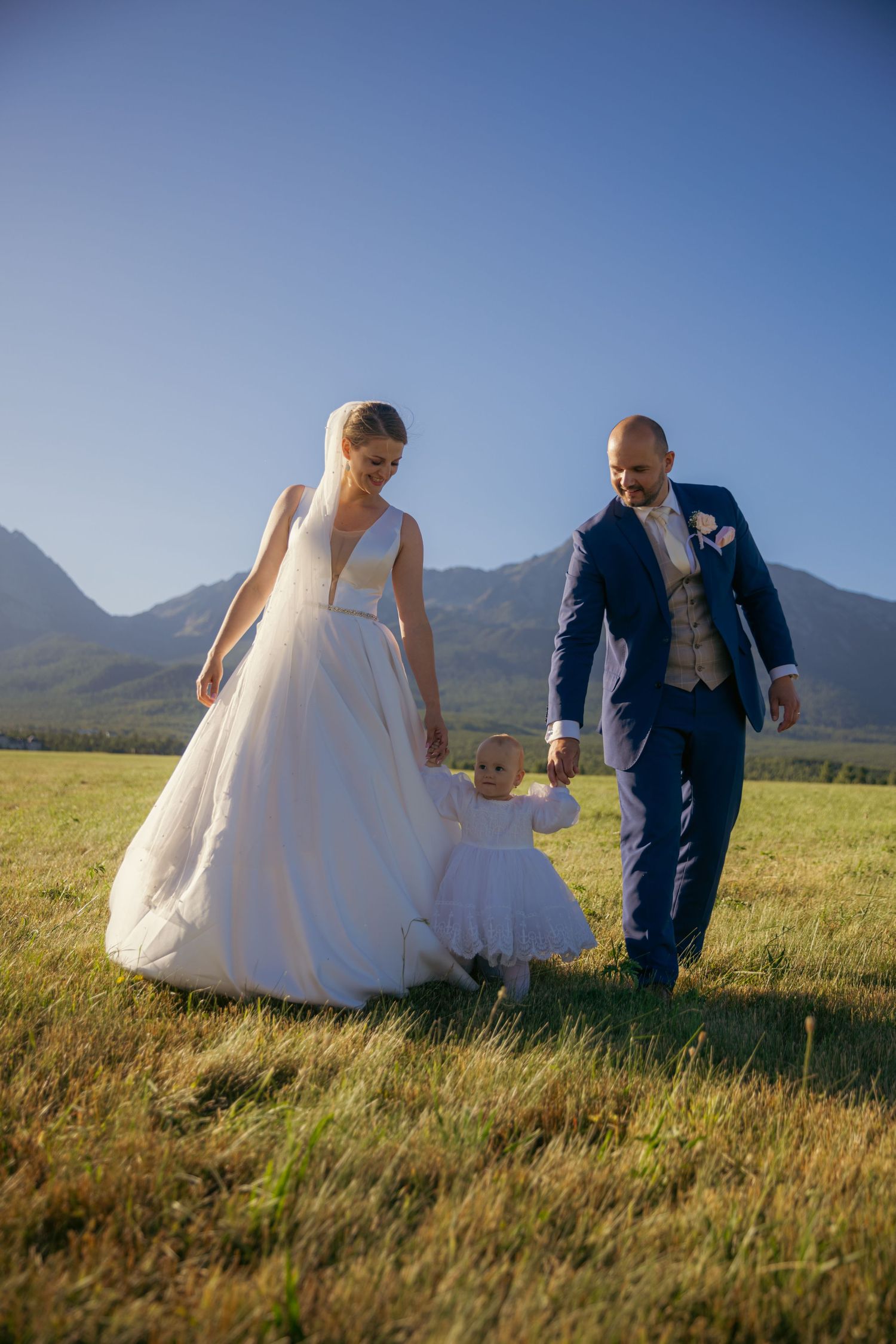 Couple in wedding attire poses playfully in a scenic mountain meadow during golden hour.