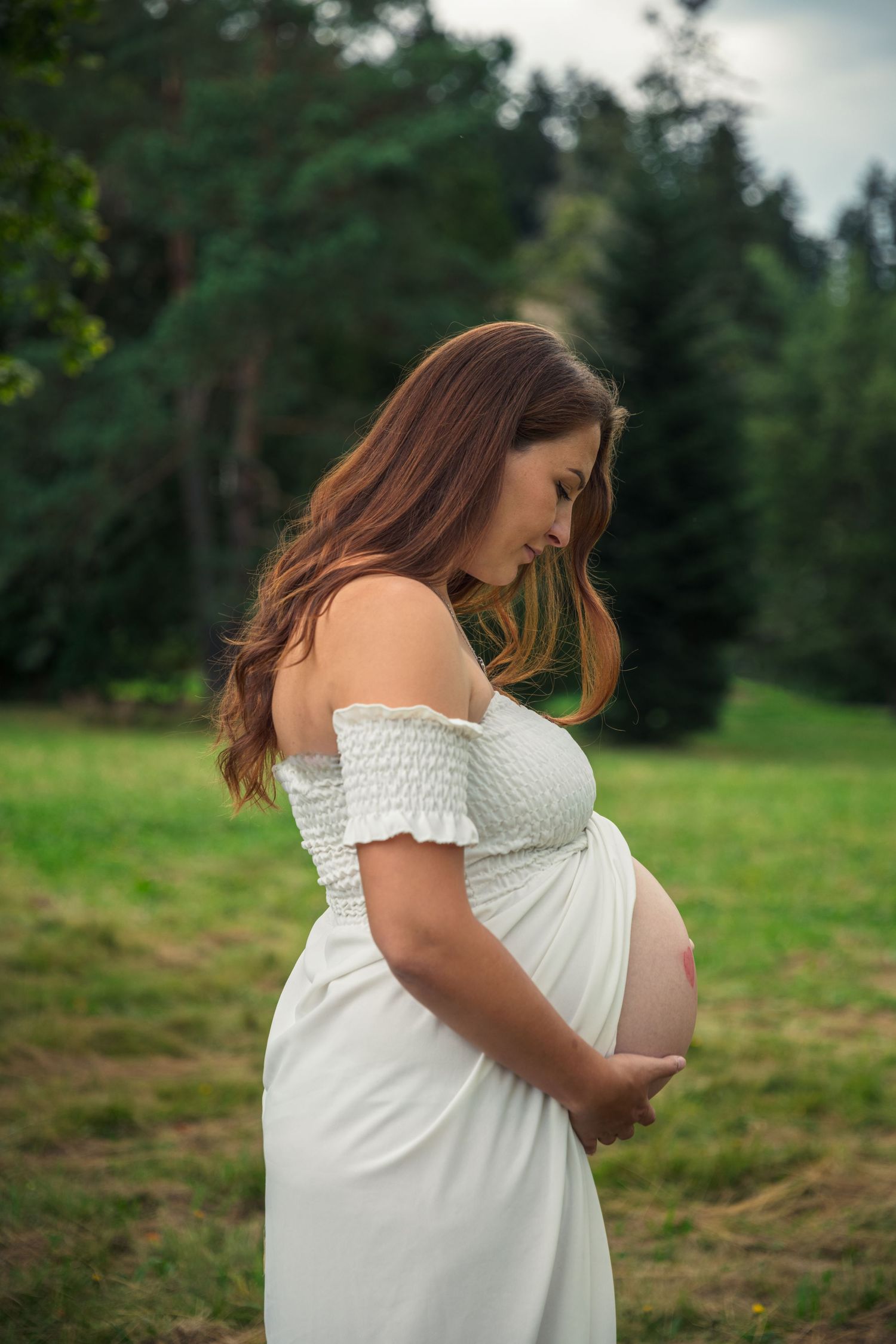 Expectant mother in white dress cradles baby bump in outdoor setting.