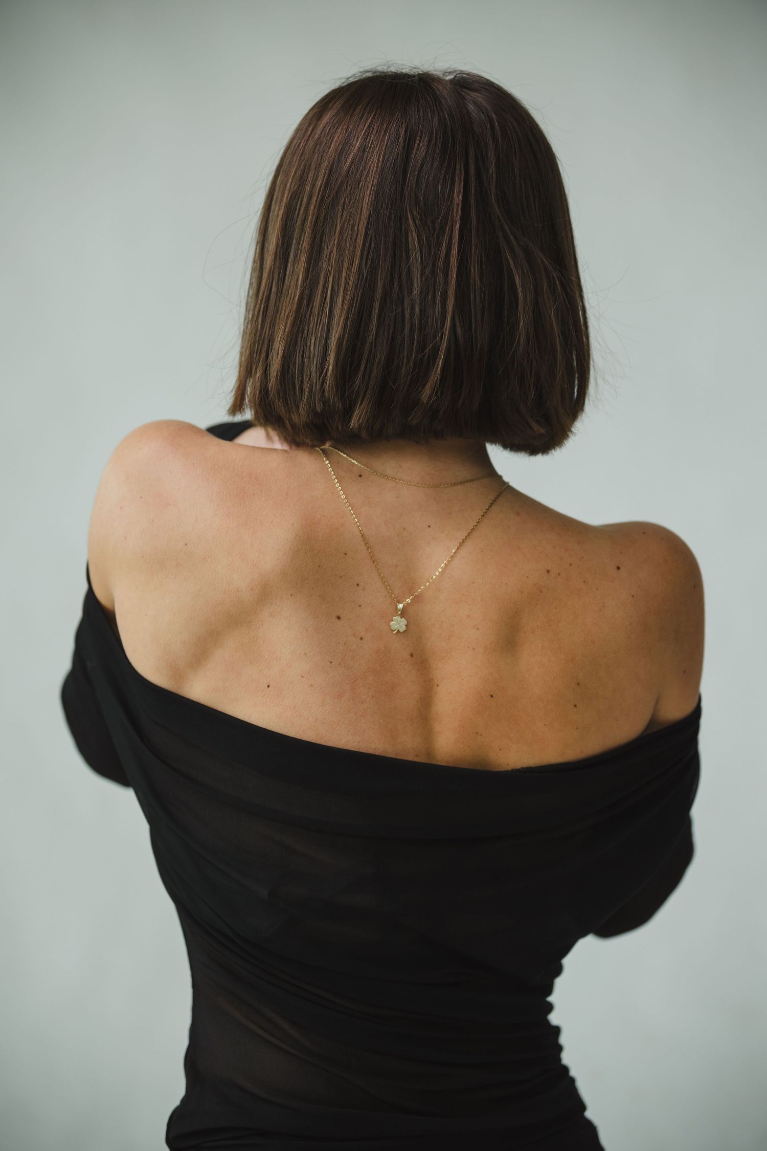 Back view of woman in black off-shoulder top showing necklace against grey background.