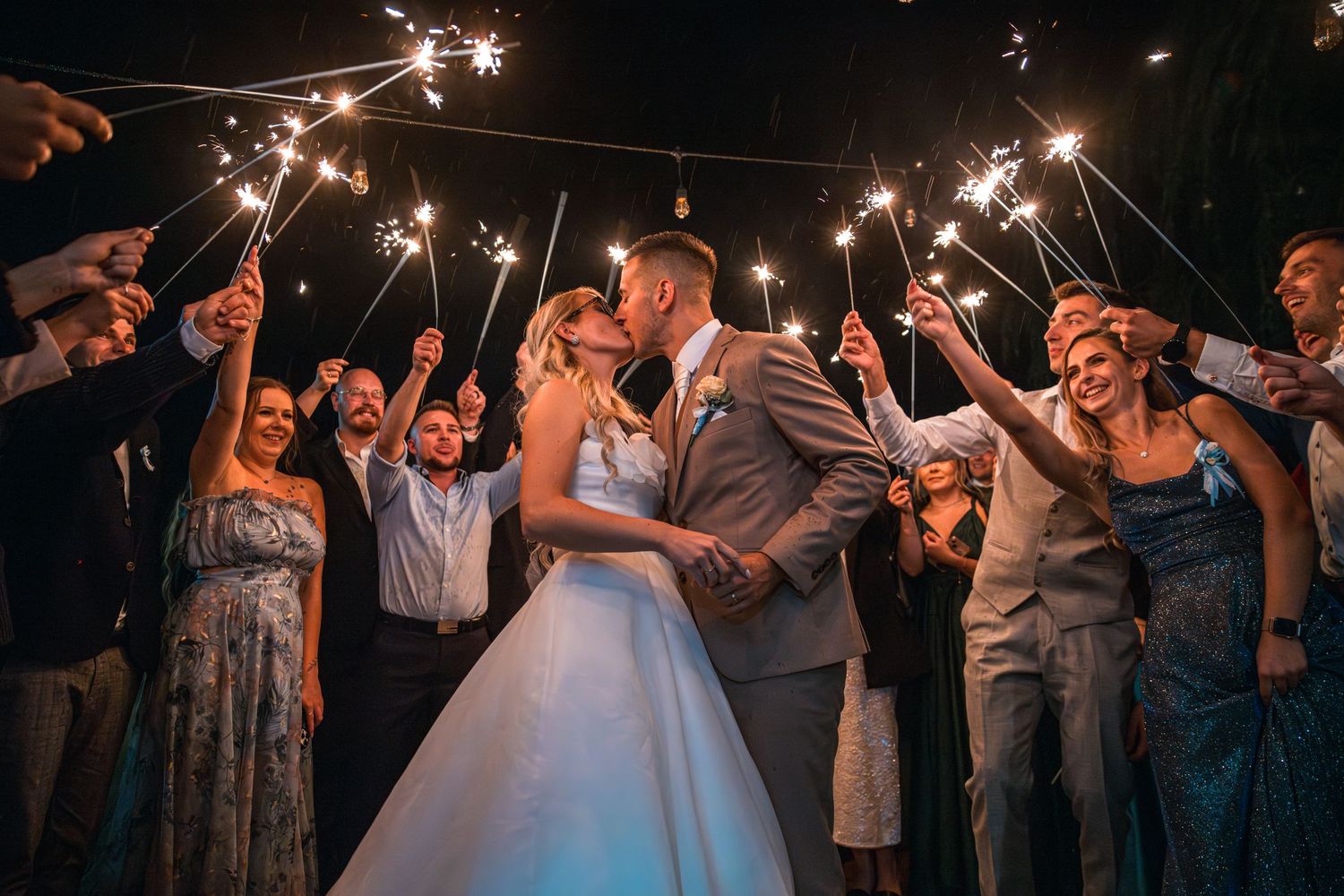 Wedding couple kisses under sparklers held by celebrating guests during evening reception.