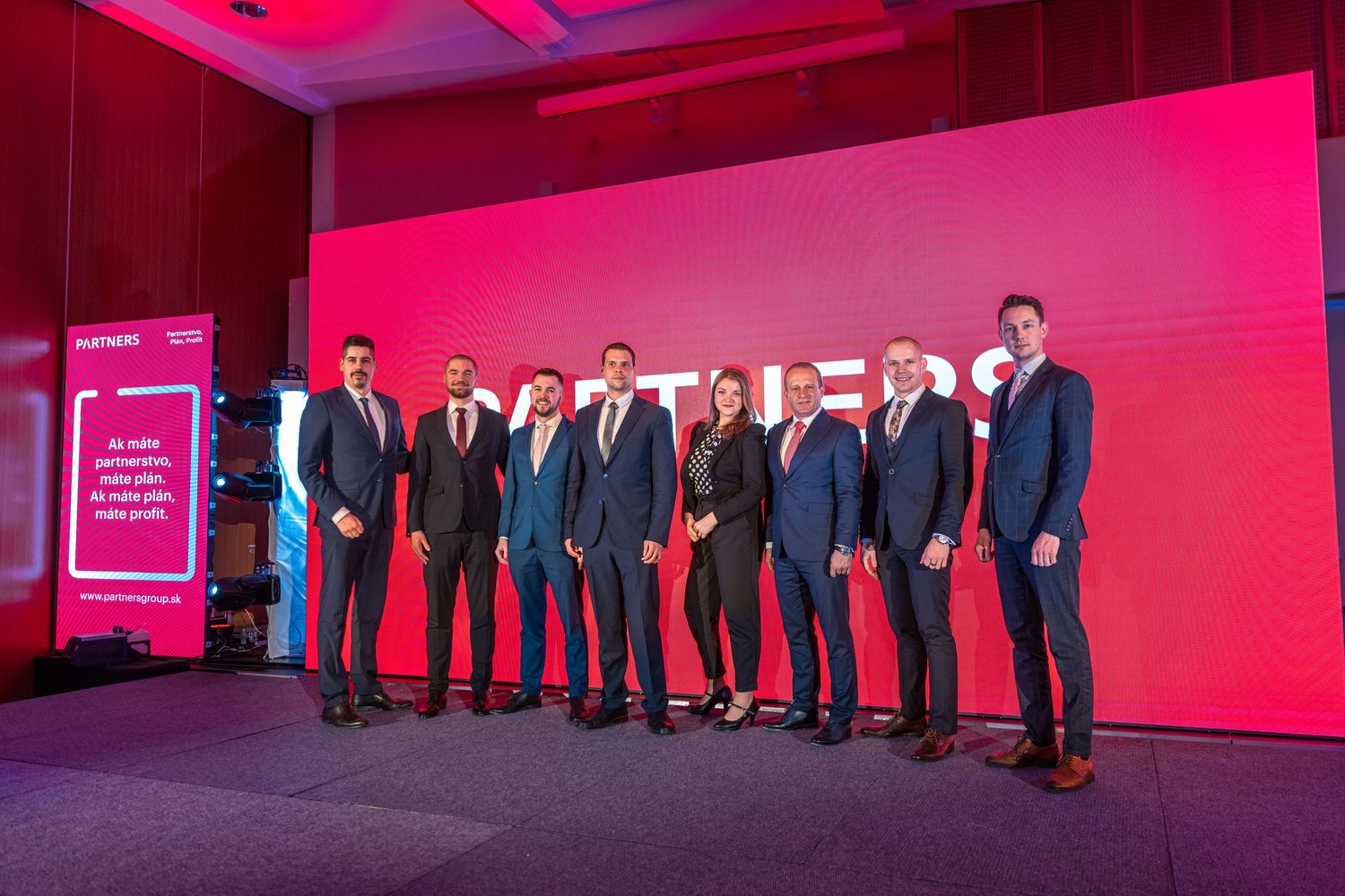 Group of business professionals lined up on stage with red backdrop at corporate event.