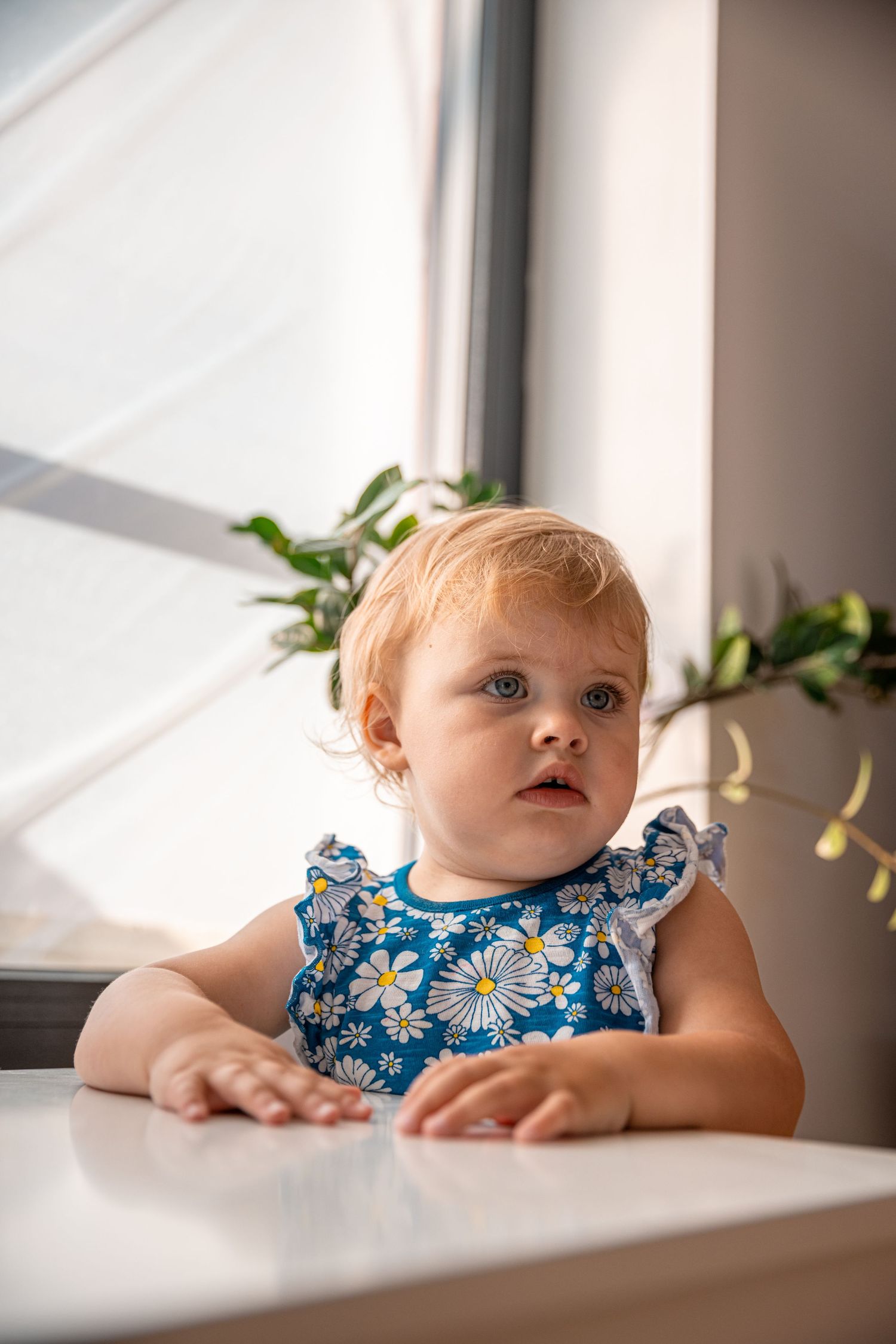 A baby in a blue floral dress looks up curiously near a sunlit window.