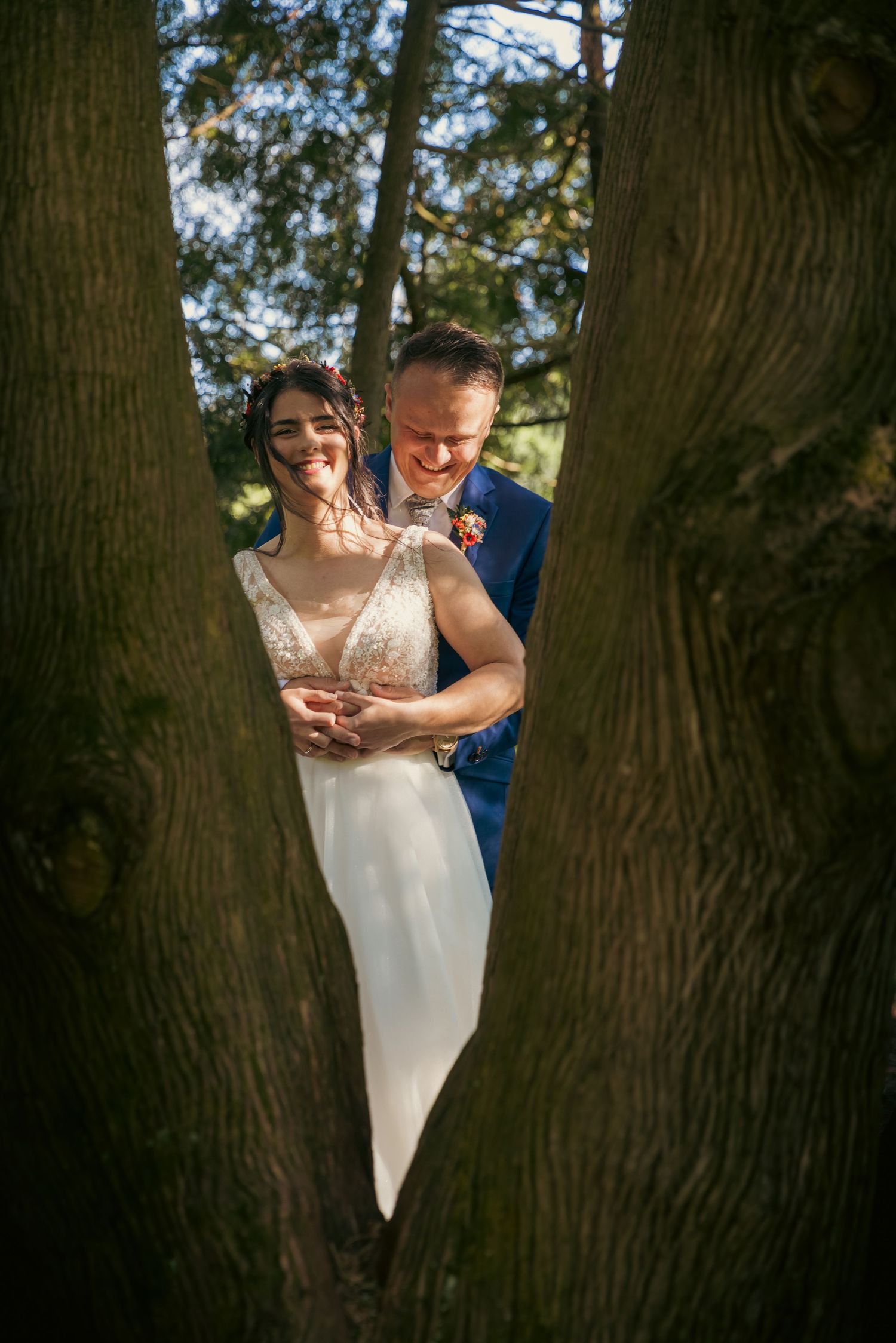 Newlyweds share an intimate moment between large tree trunks in a forest setting.