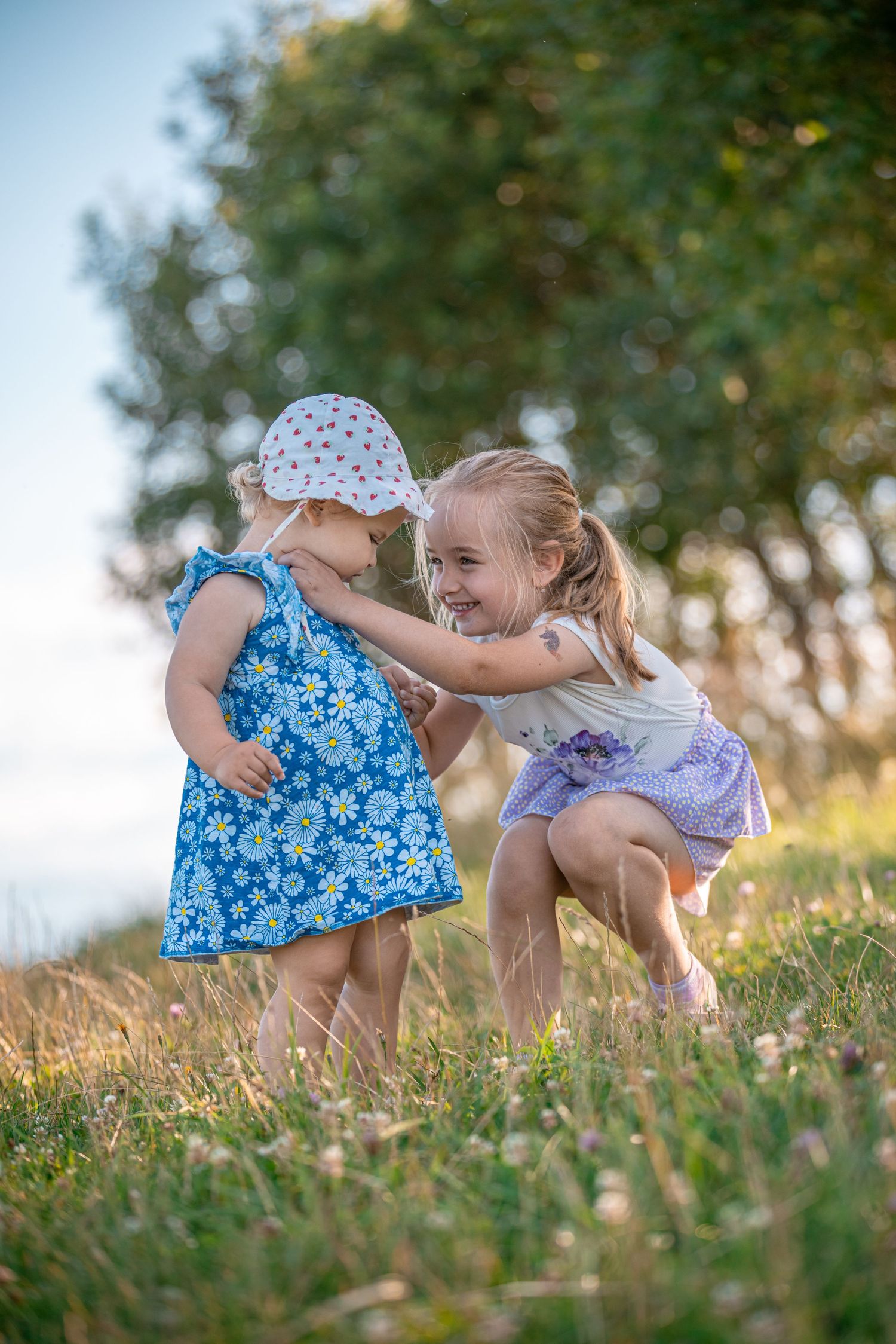 Two children in floral dresses play together in a grassy field at golden hour.