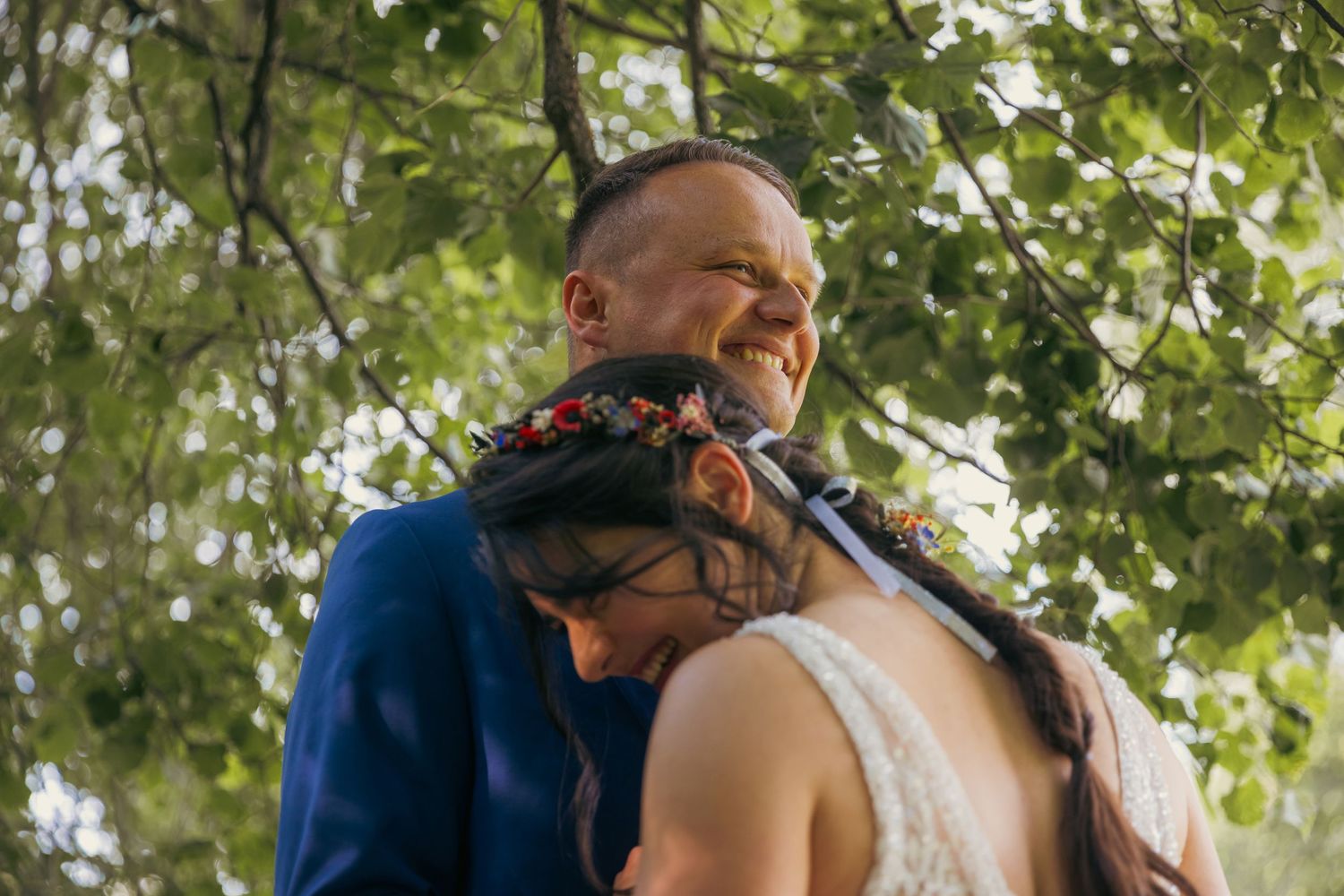 Wedding couple embraces among green leafy trees in natural outdoor setting.