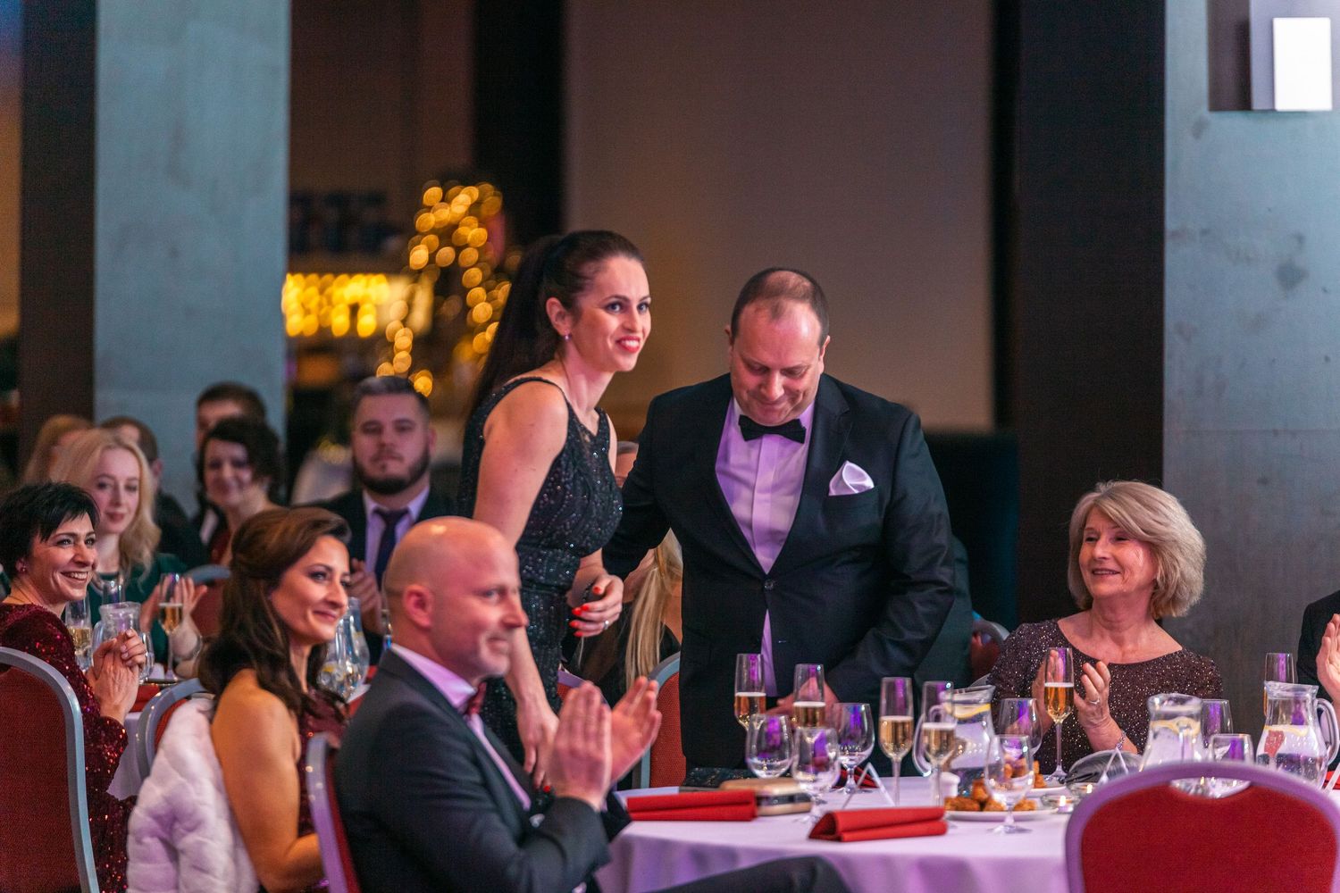 Elegant formal event with guests seated at round tables in a dimly lit banquet hall with red and purple lighting.