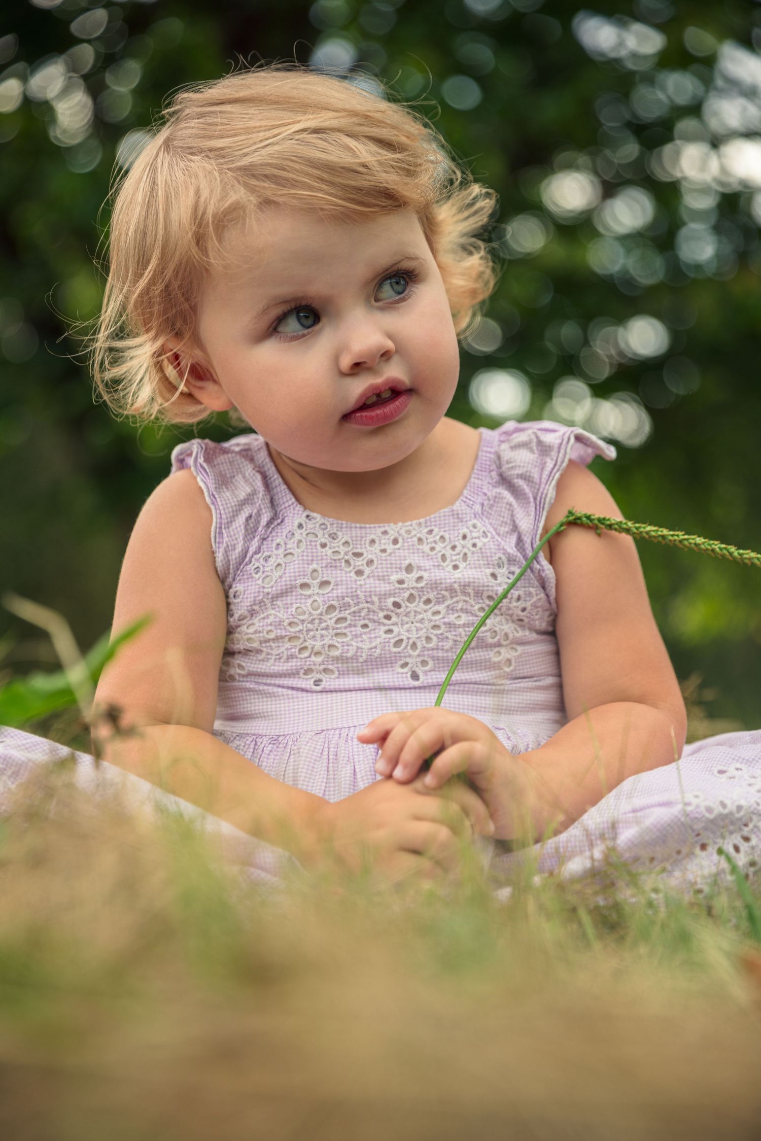 Young child in lavender dress sits in grass with dreamy expression.