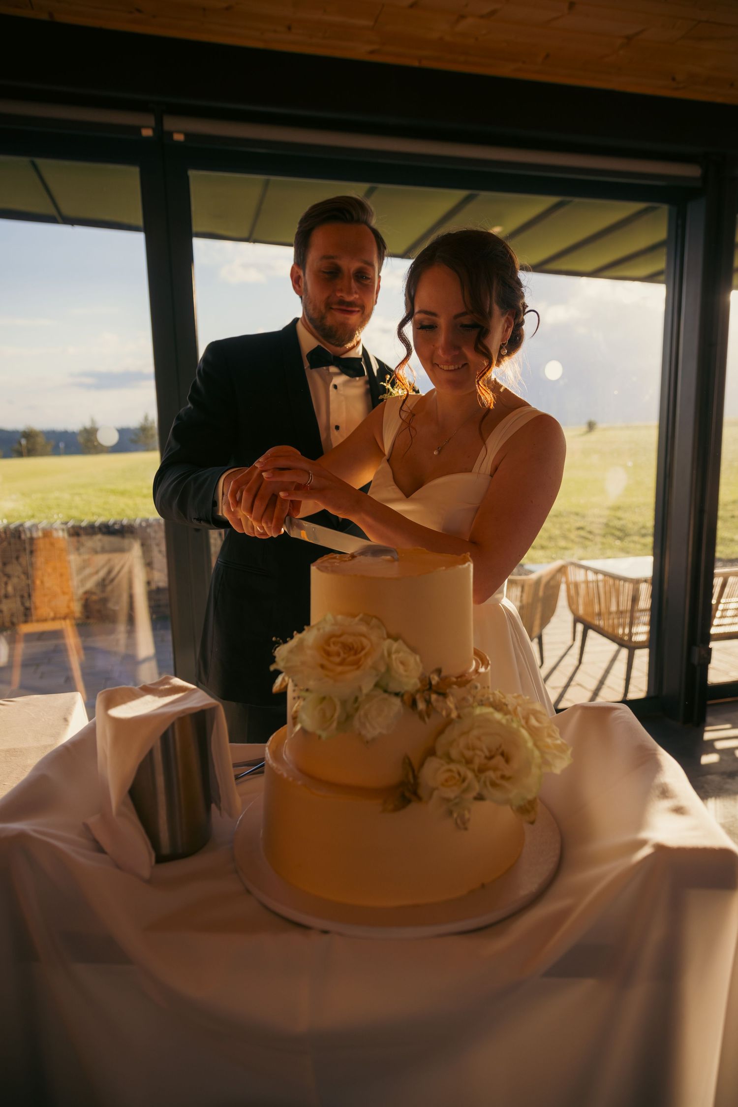 Newlyweds cut their elegant white wedding cake together at an indoor reception venue.