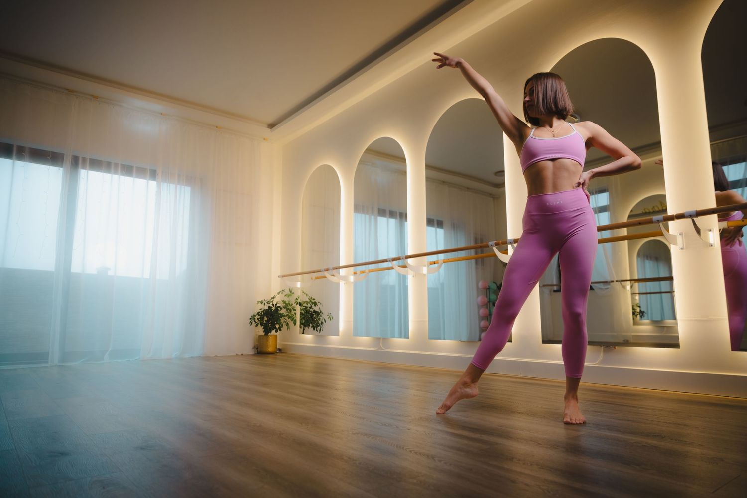 Woman in pink workout attire practicing dance moves in a sunlit studio with ballet barre.
