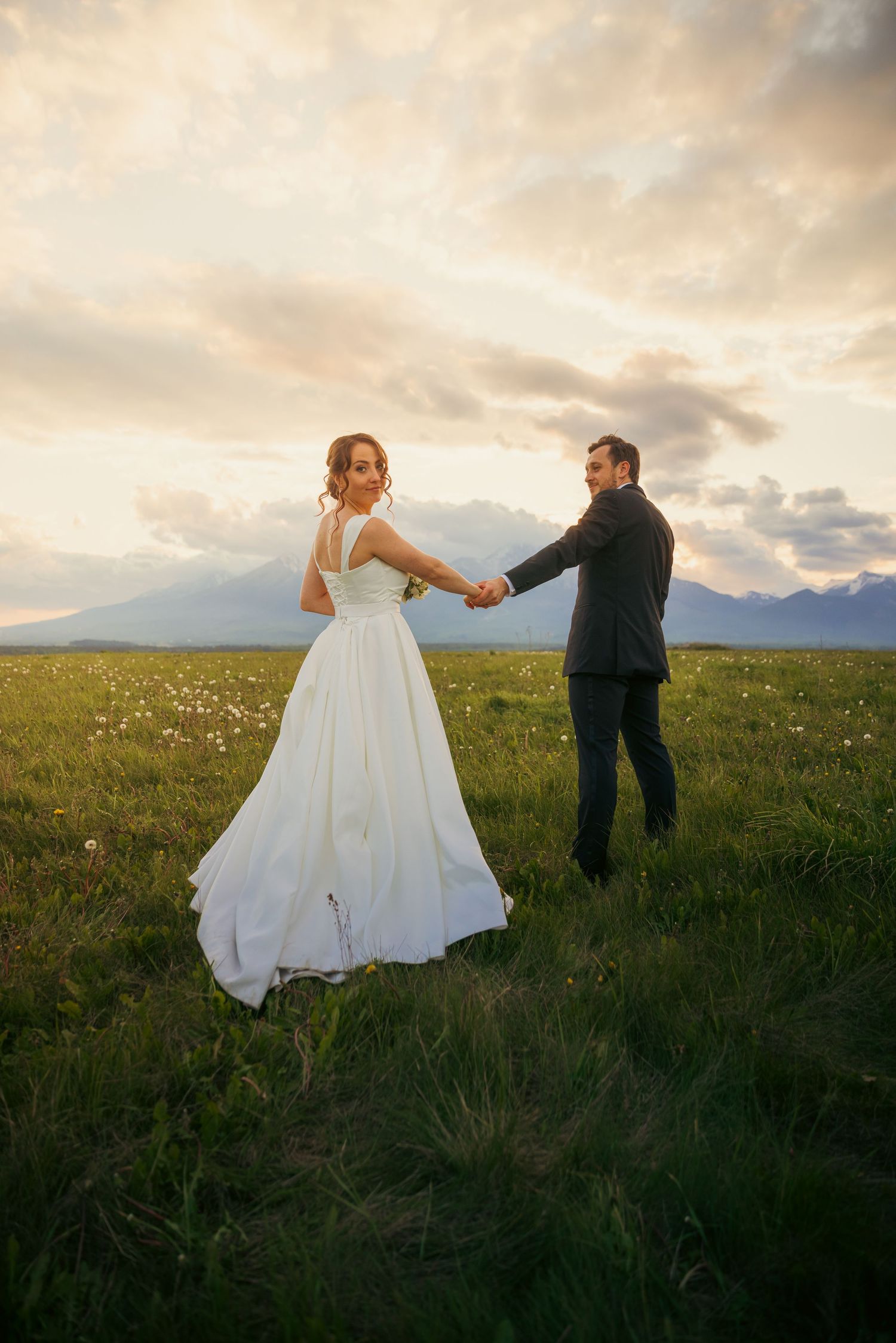 Romantic sunset wedding photo in mountain meadow with couple holding hands.