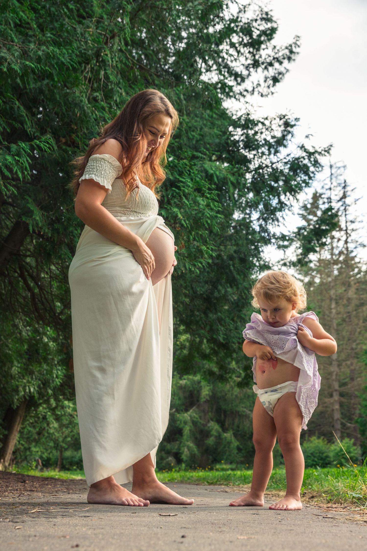 Pregnant woman in white dress stands with toddler on outdoor path.