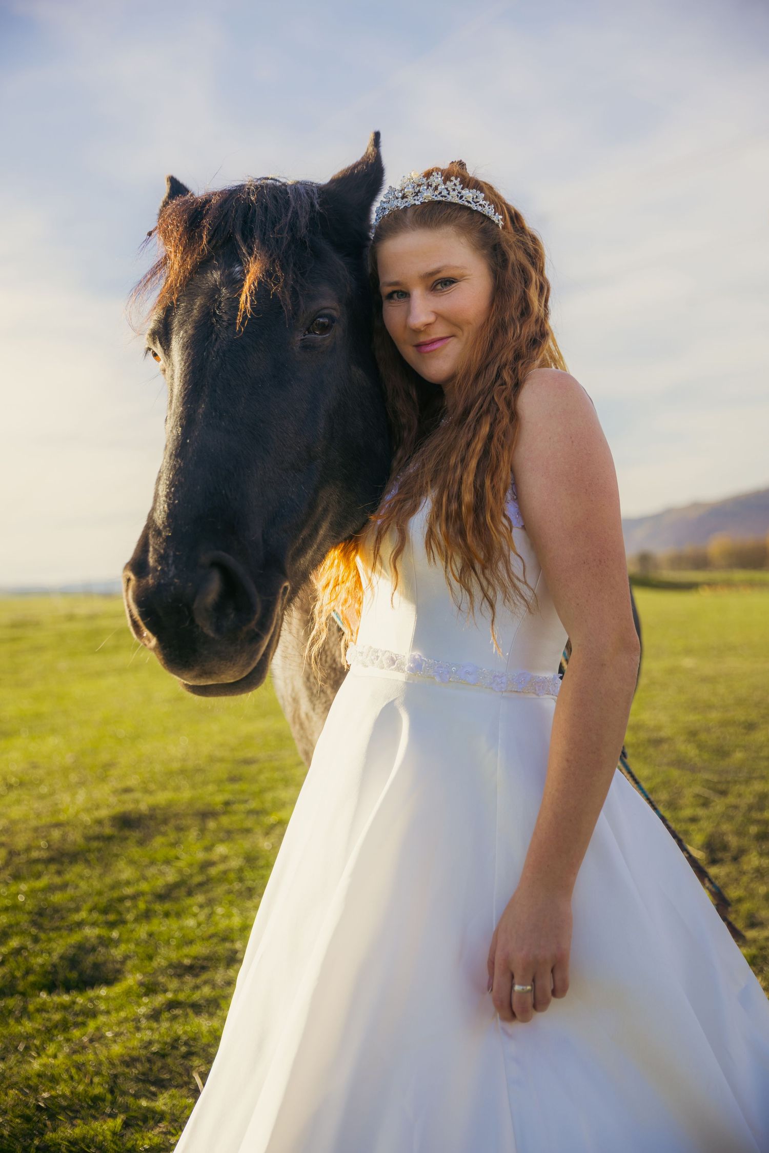 A bride in a white gown poses with a black horse in a sunlit meadow with mountains in the background.