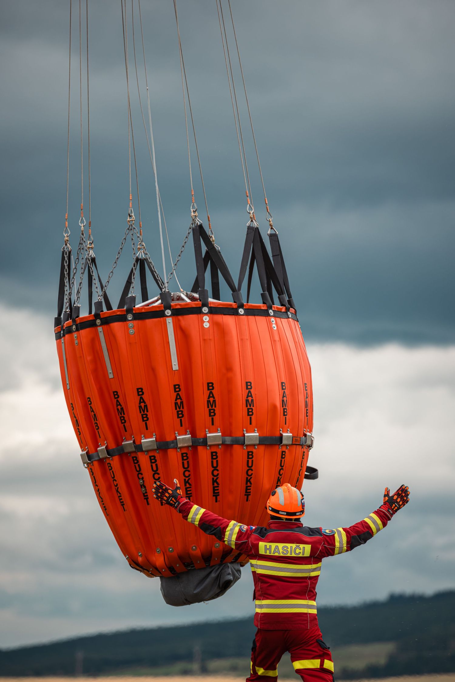 Firefighters guide a large orange water bucket suspended from helicopter cables during aerial operations.
