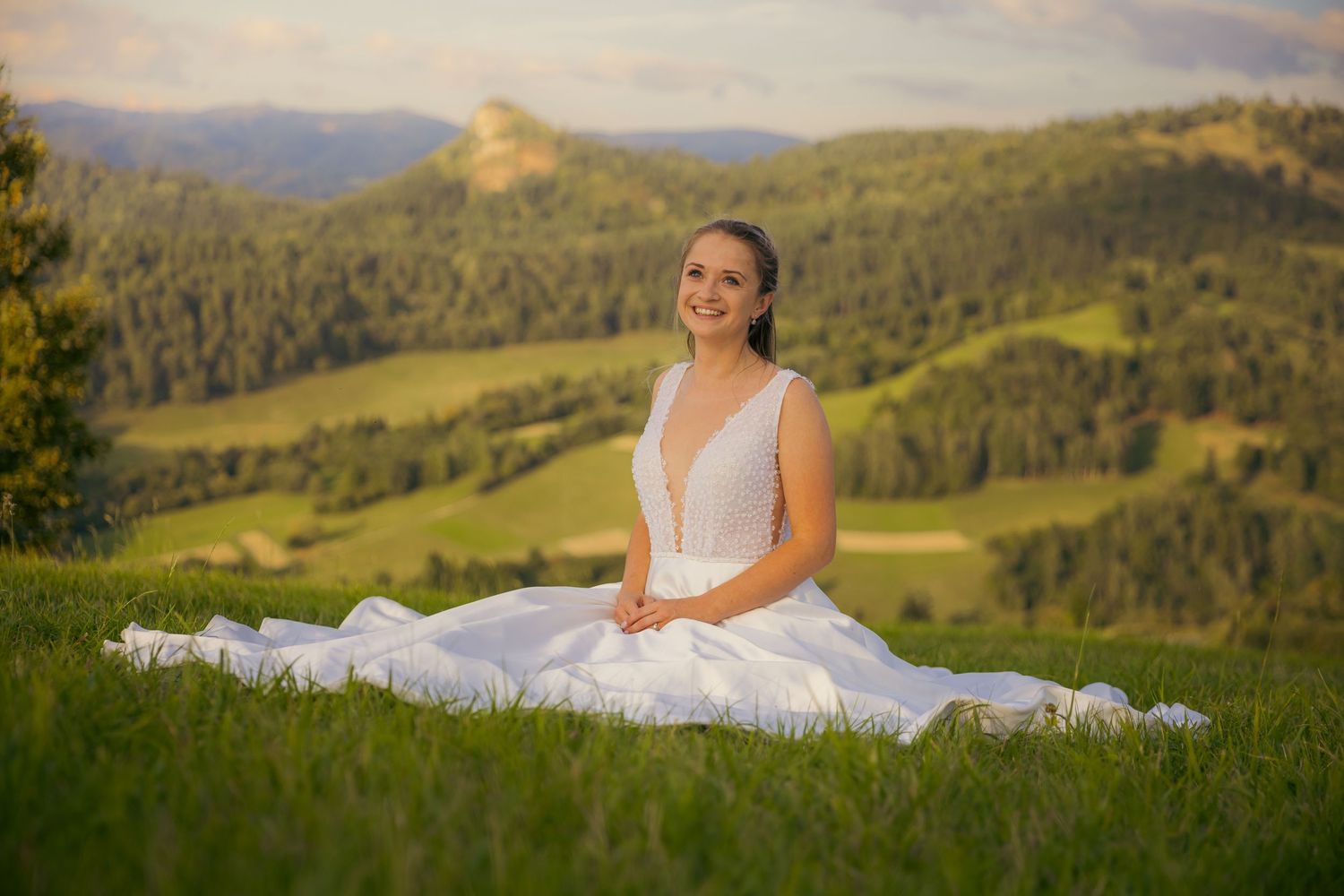 A wedding dress flows elegantly across rolling green hills with mountains in the background at sunset.