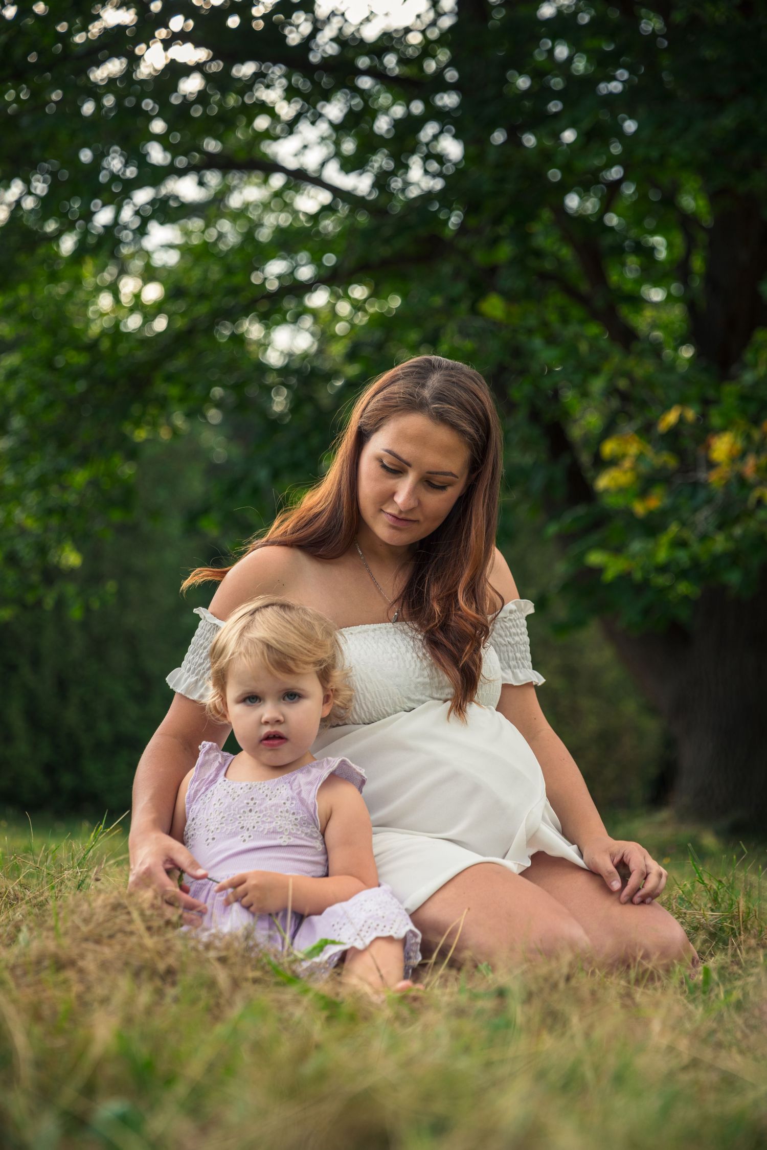 Mother and child sit together on grassy hill during outdoor photoshoot.