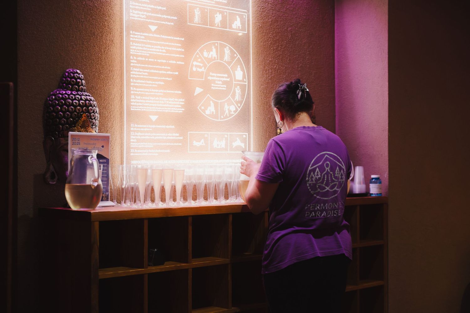 Person in purple shirt working at illuminated bar station with glassware display.
