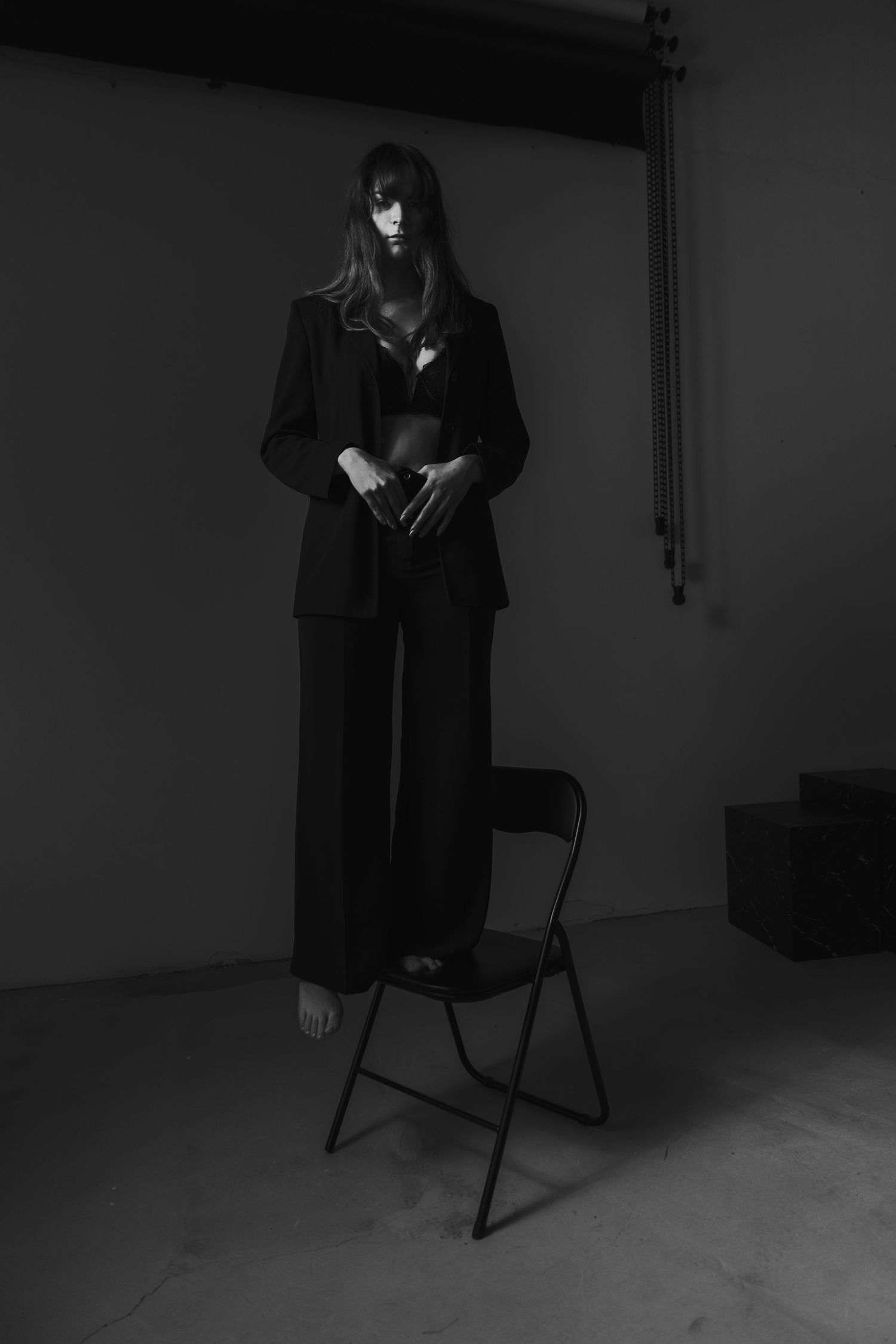 Figure in black suit stands on folding chair in dramatic monochrome portrait.