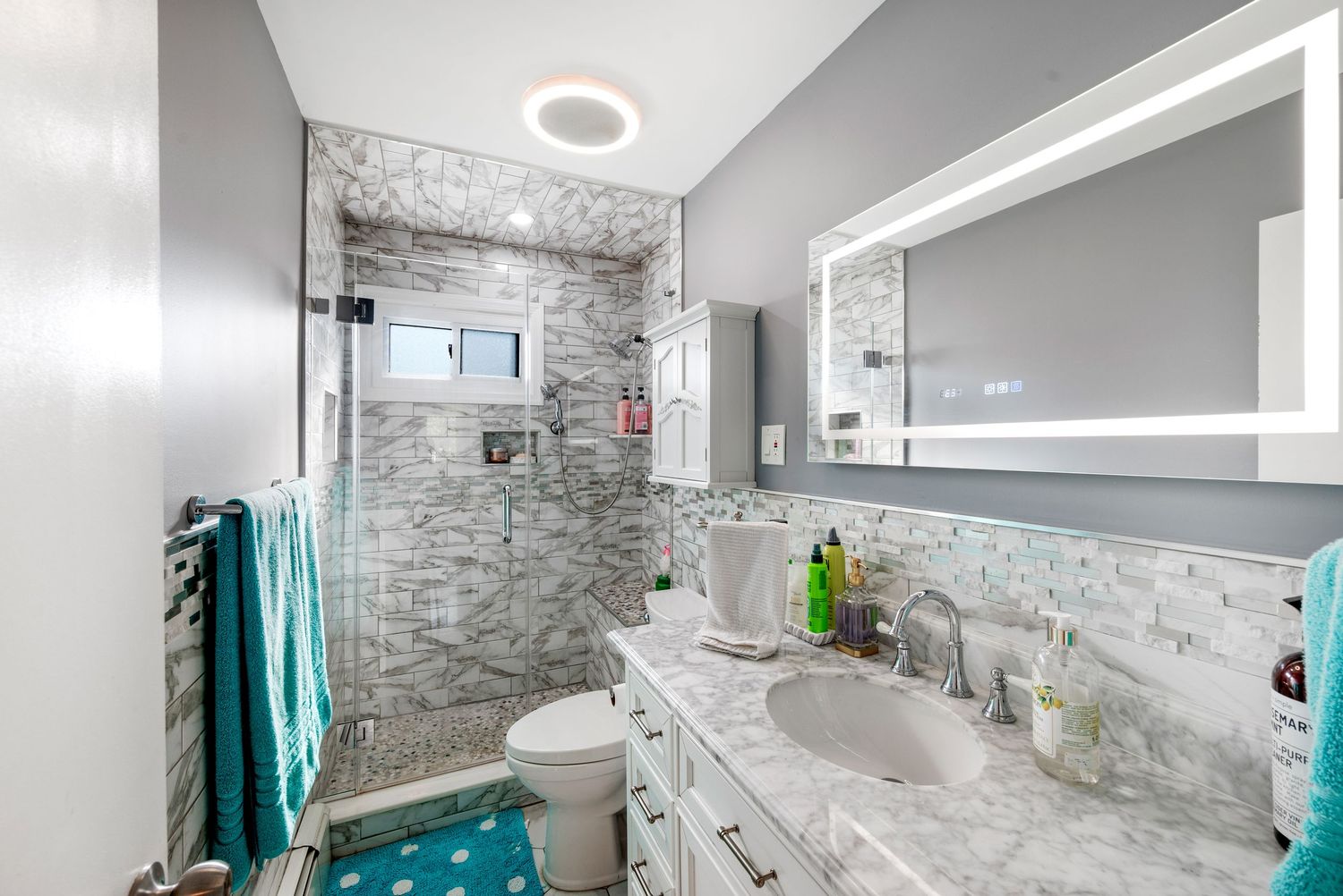 Renovated bathrooom with grey walls featuring a walk-in shower, a large mirror, white fixtures, and matching blue decor.