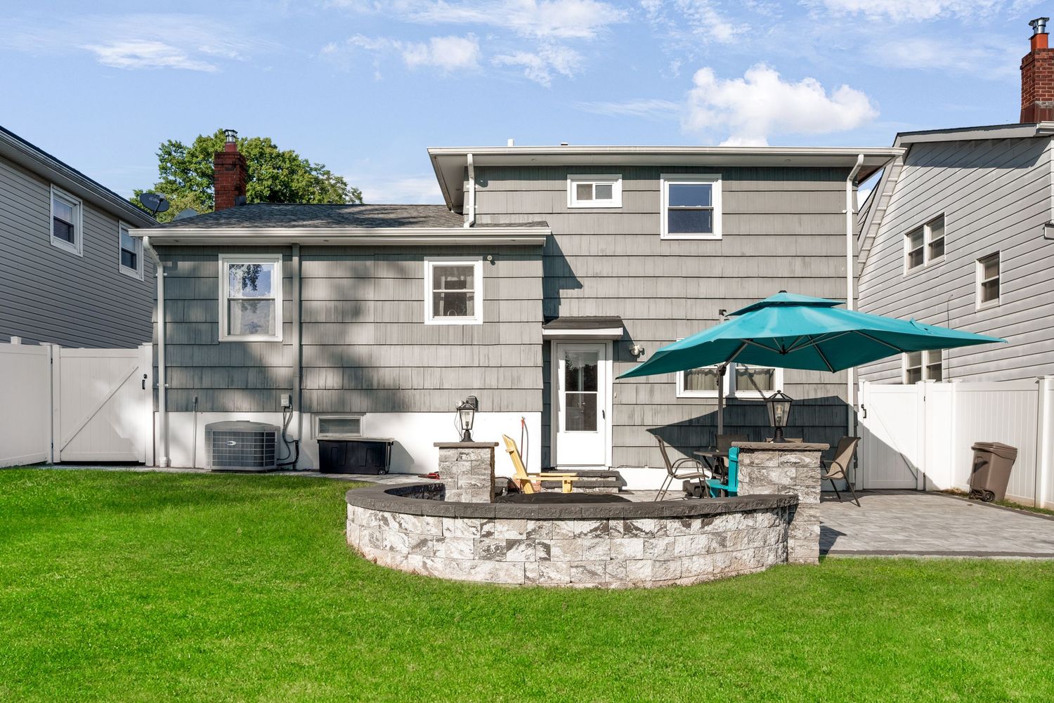 Exterior of back of grey split-level house featuring green grass, blue sky, a stone patio with blue umbrella and patio furniture.