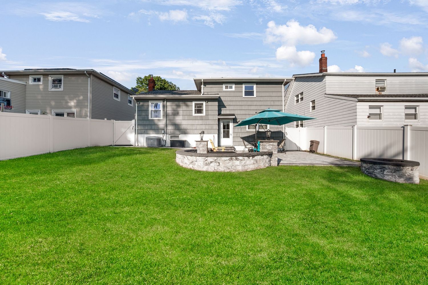 Exterior of back of grey split-level house featuring green grass, blue sky, a stone patio with blue umbrella and patio furniture.