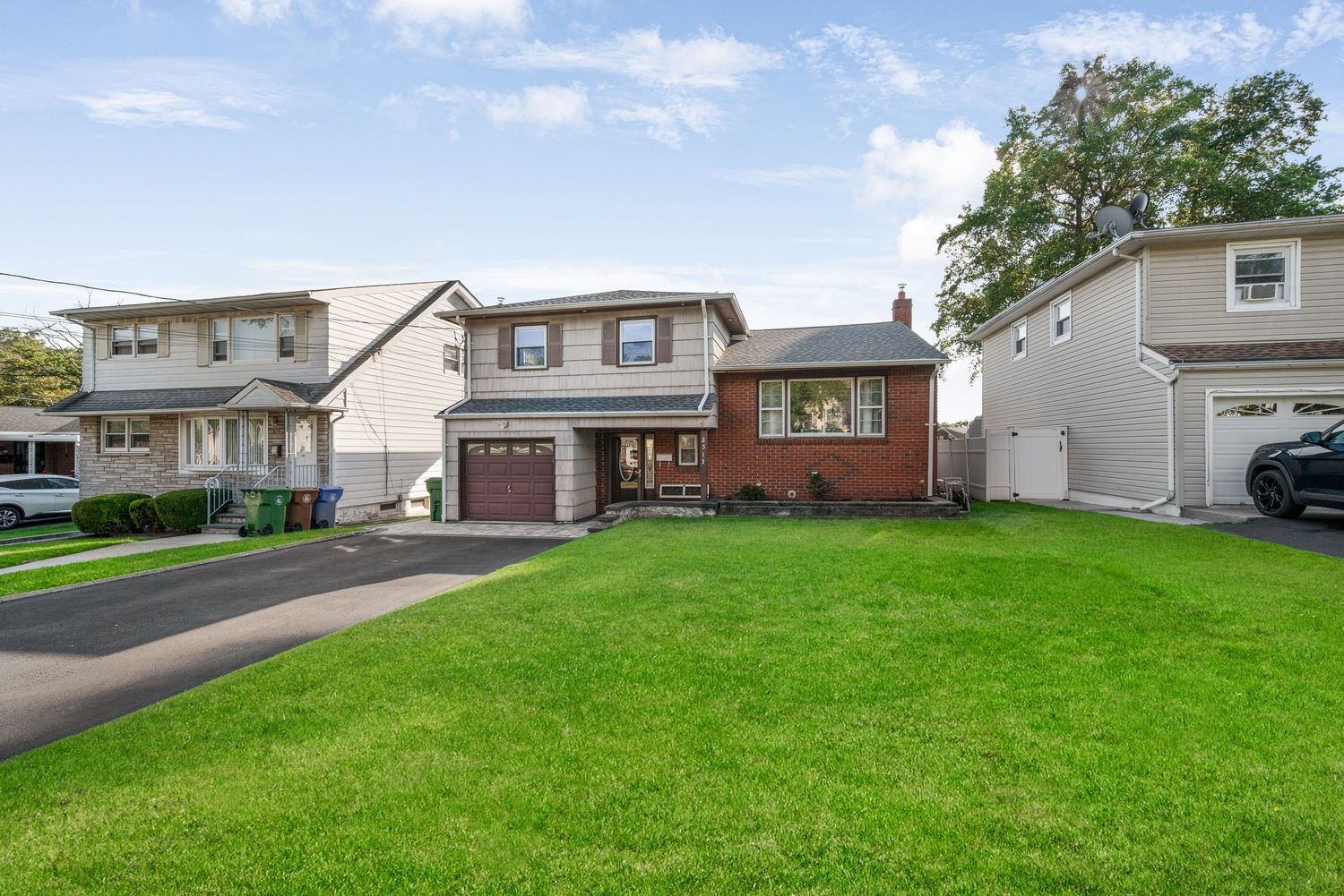 Exterior street level front of split-level home with grey siding and red brick, garage, double asphalt driveway, green grass, and blue sky
