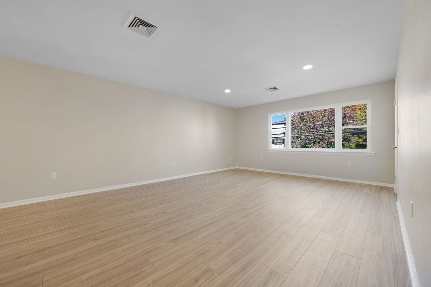 Large living room with light hardwood floors and beige walls featuring a large window.