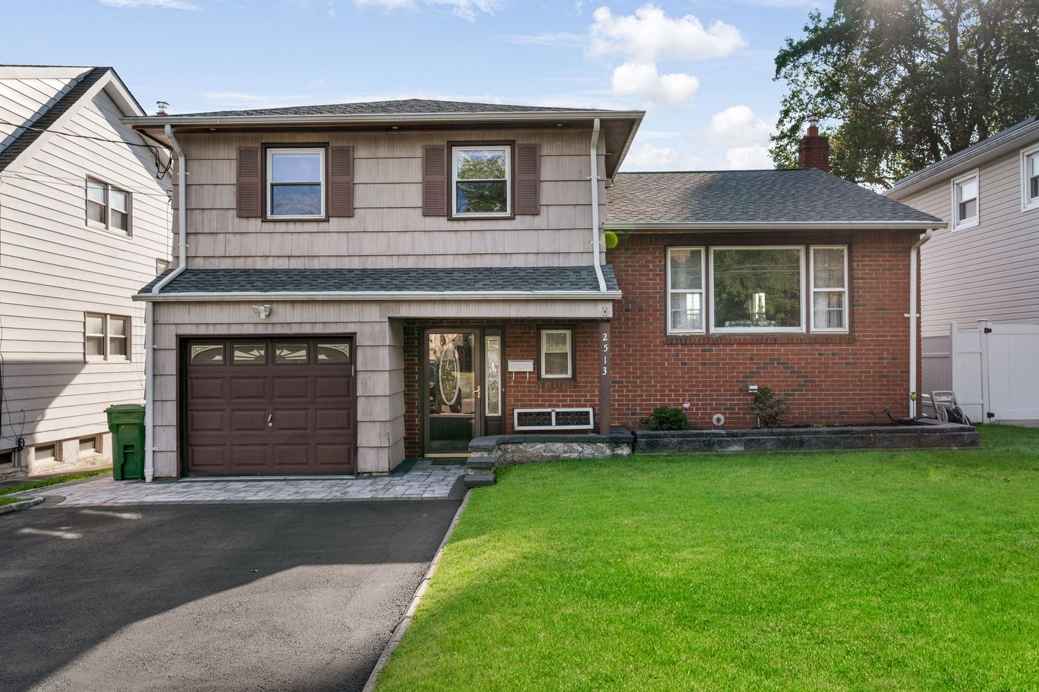 Exterior front of split-level home with grey siding and red brick, garage, double asphalt driveway, green grass, and blue sky