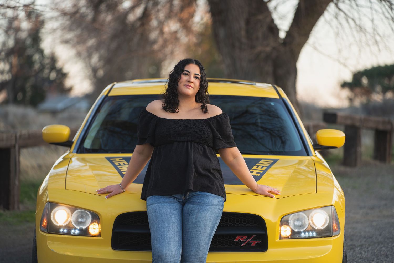 Young woman in black top and jeans posing confidently in front of a bright yellow Dodge Charger outdoors.