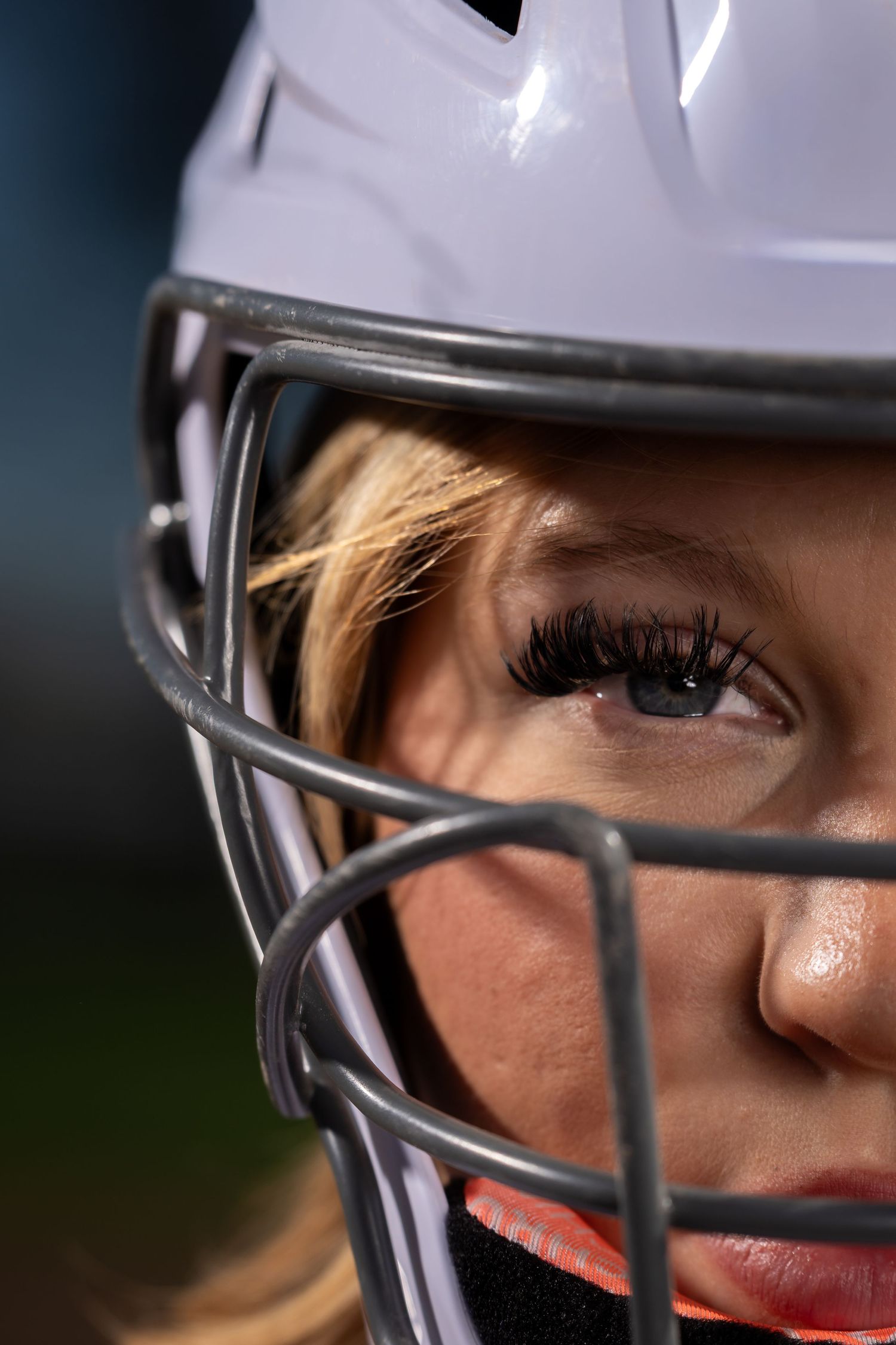 Close-up of a female softball player wearing a white batting helmet with face guard, intense focused expression.