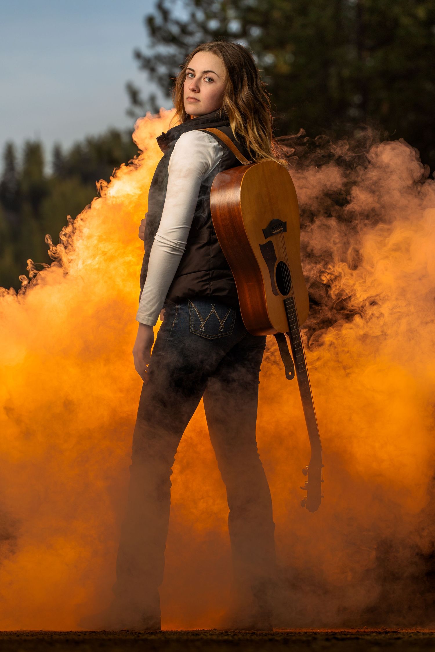 Young woman with acoustic guitar poses dramatically in orange smoke clouds outdoors.