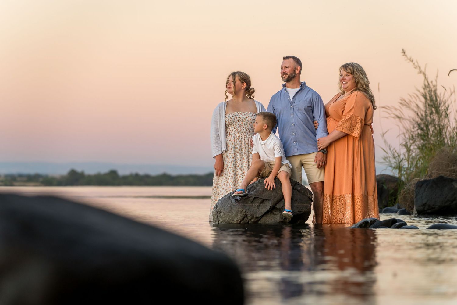 Family standing in shallow water at sunset wearing casual clothing against a warm sky backdrop.