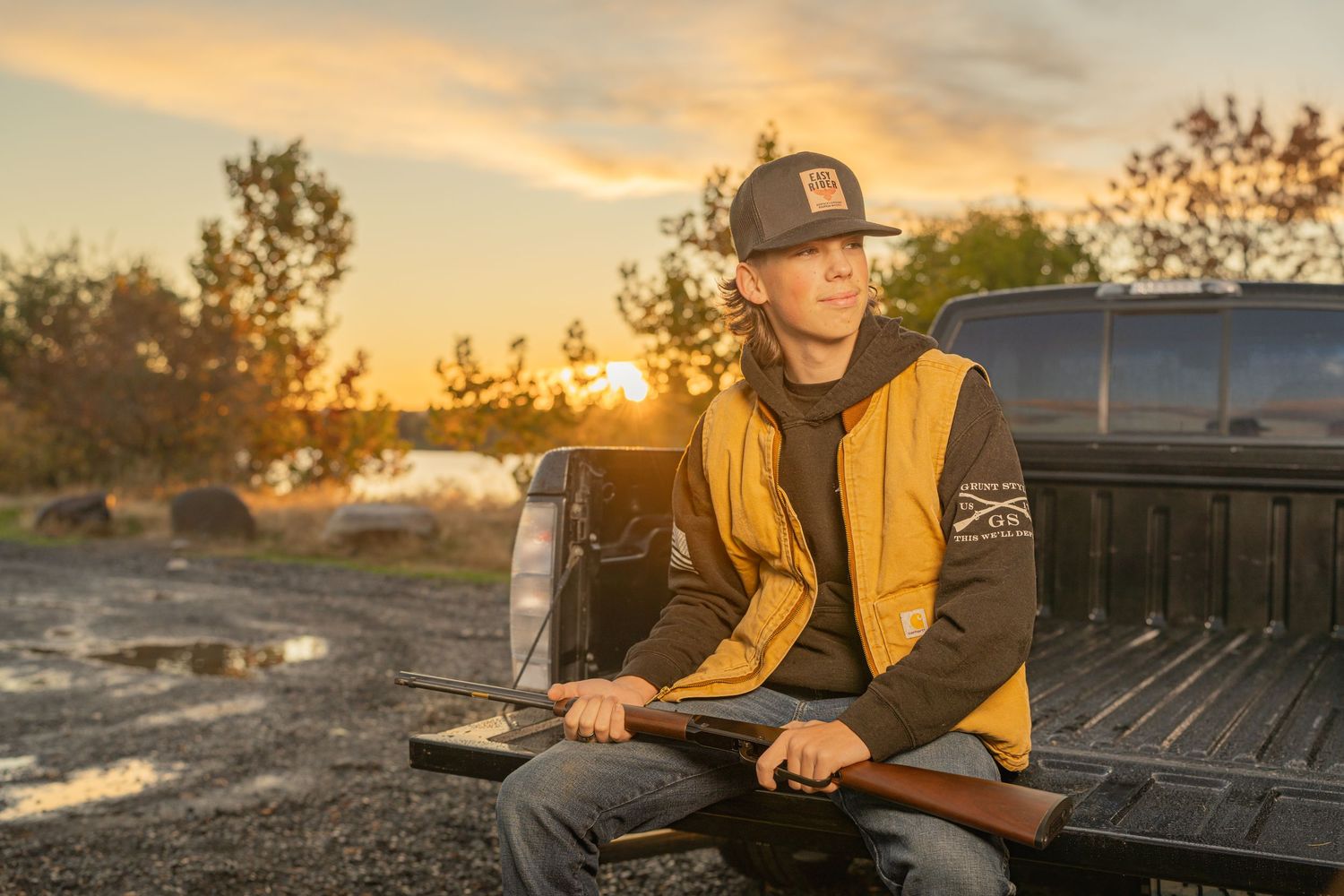 Someone sits in the bed of a pickup truck at sunset wearing a yellow and black jacket and baseball cap.