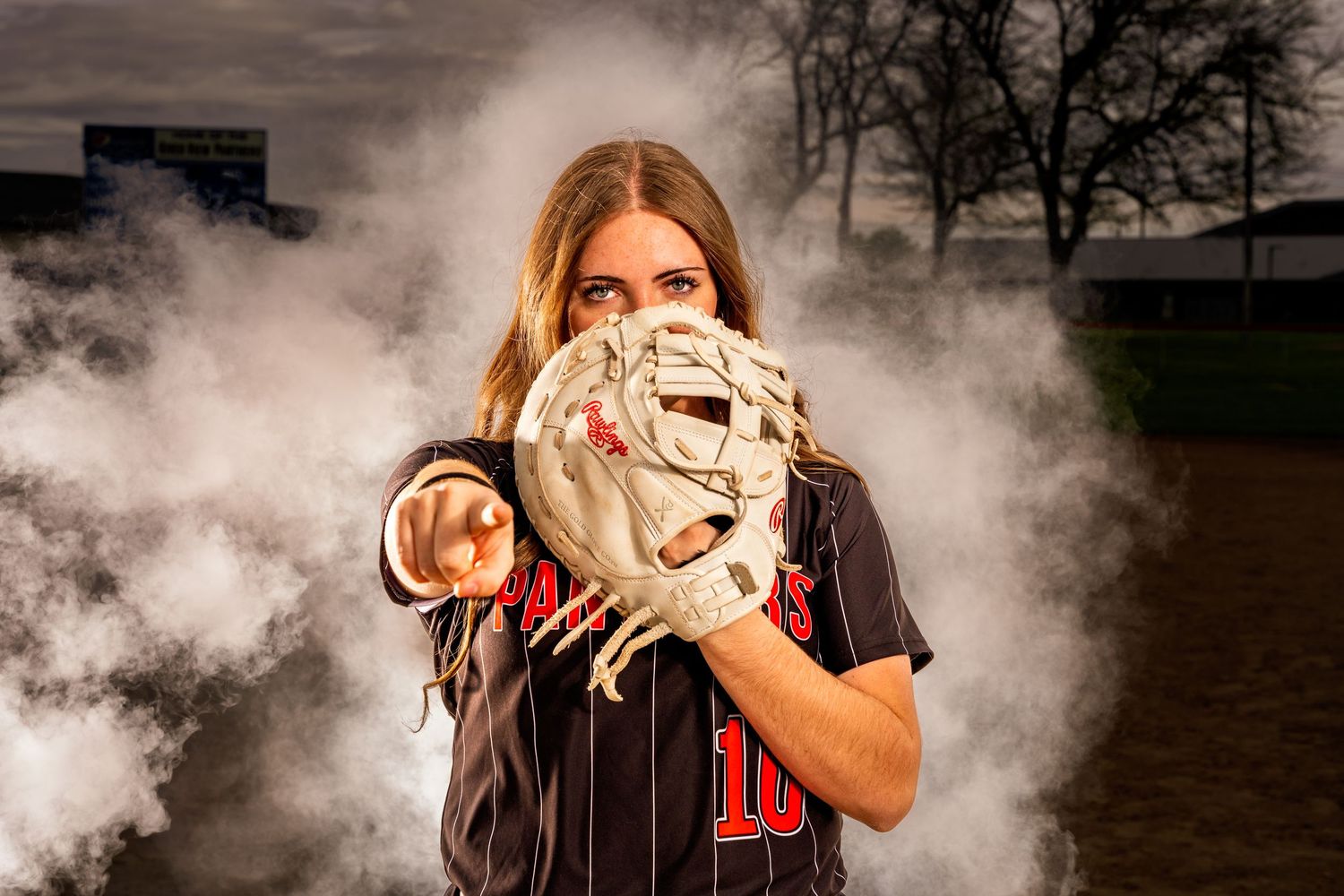 Softball player poses with glove and dramatic smoke effects in outdoor sports photoshoot.
