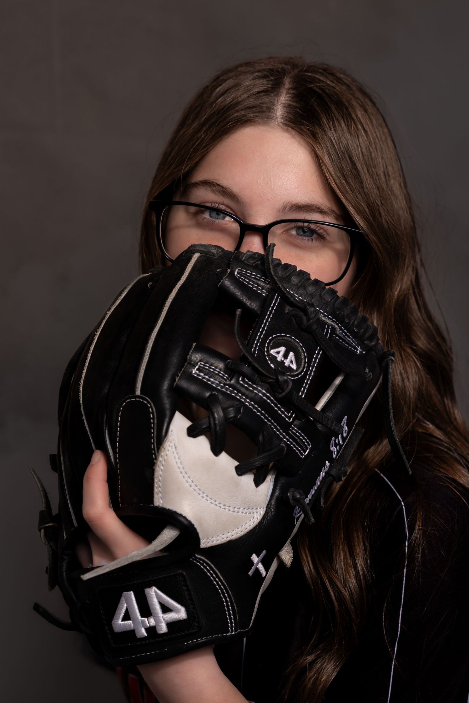 Young girl with glasses holding a black and white 44 brand baseball glove up to her face in a studio portrait.