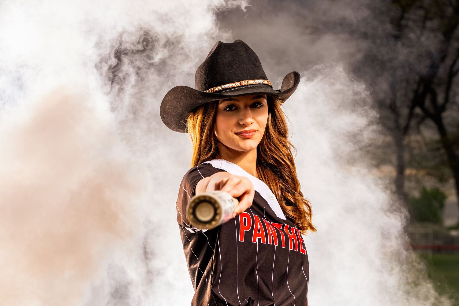 Softball player poses in cowboy hat with dramatic smoke effects in outdoor setting.