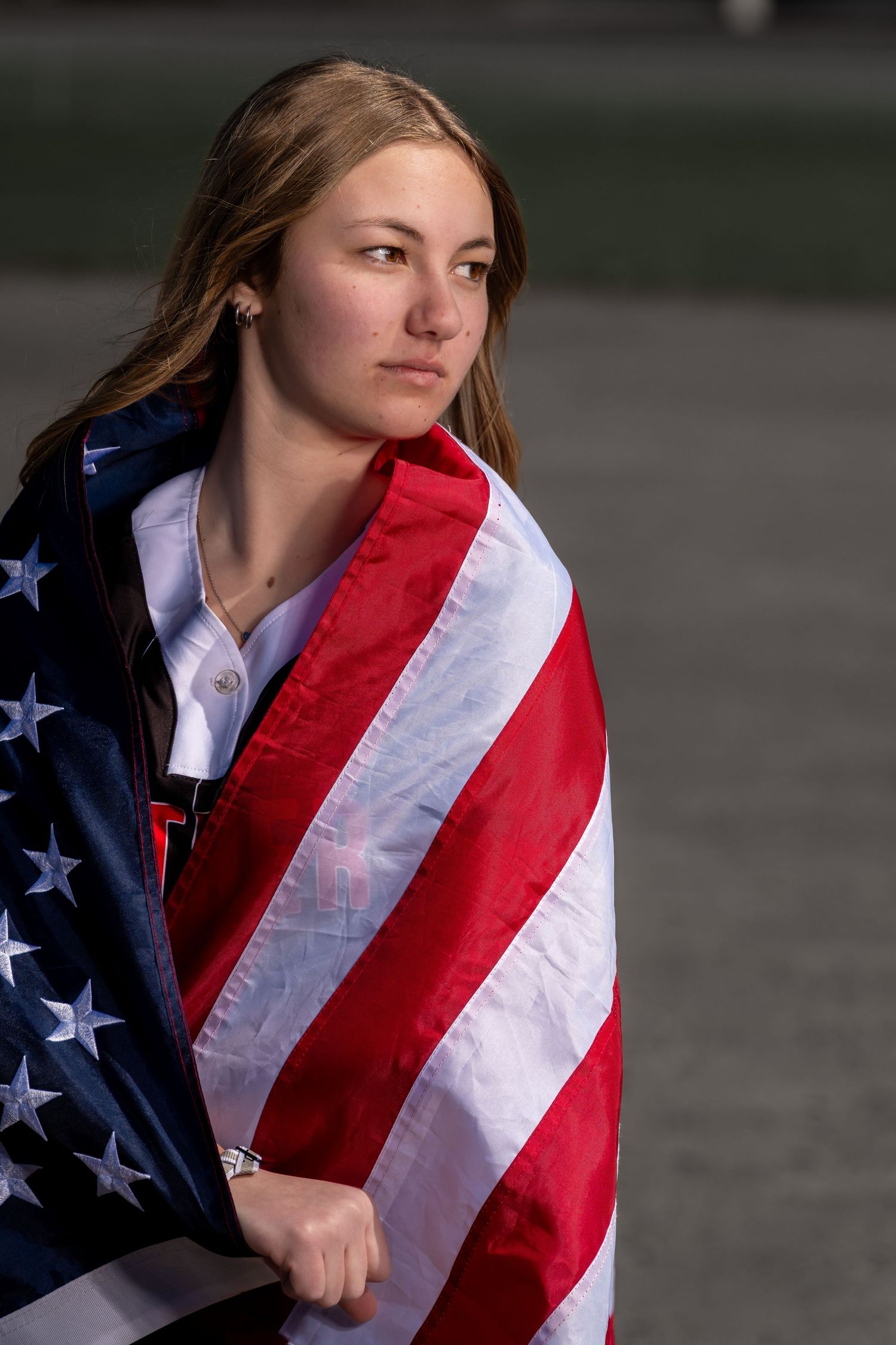 Young woman wrapped in an American flag, gazing thoughtfully into the distance outdoors.