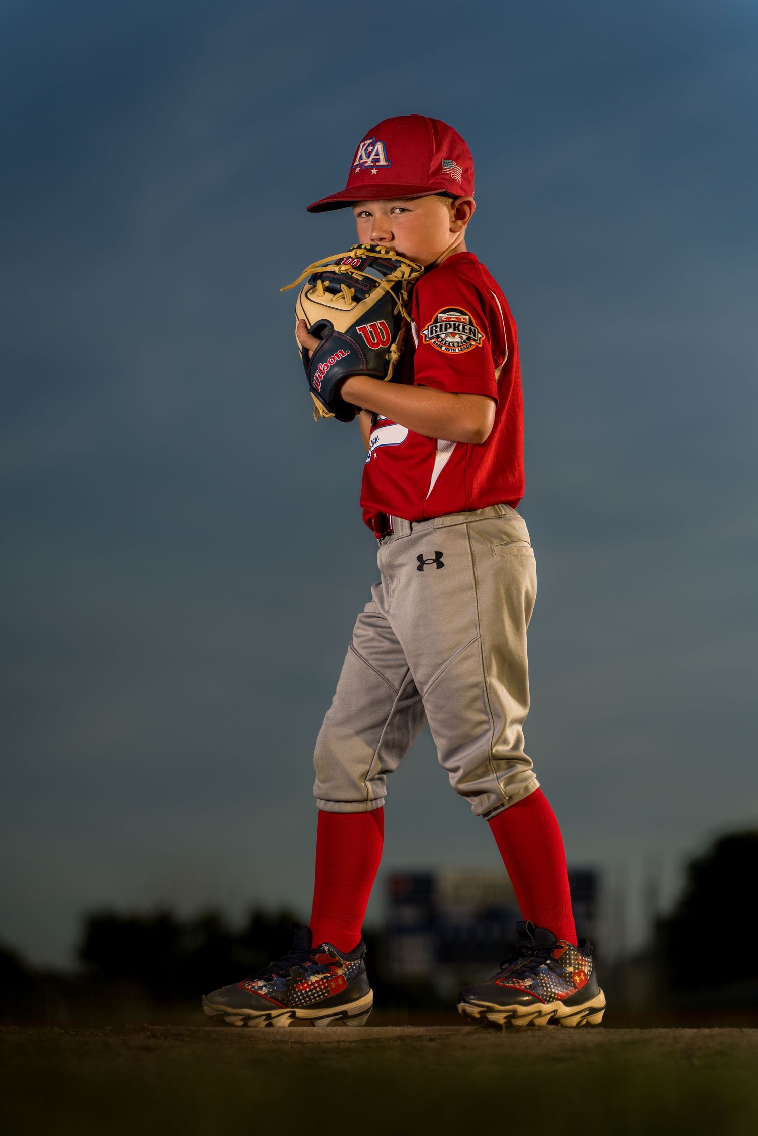 Young baseball player in uniform stands ready on field at dusk.