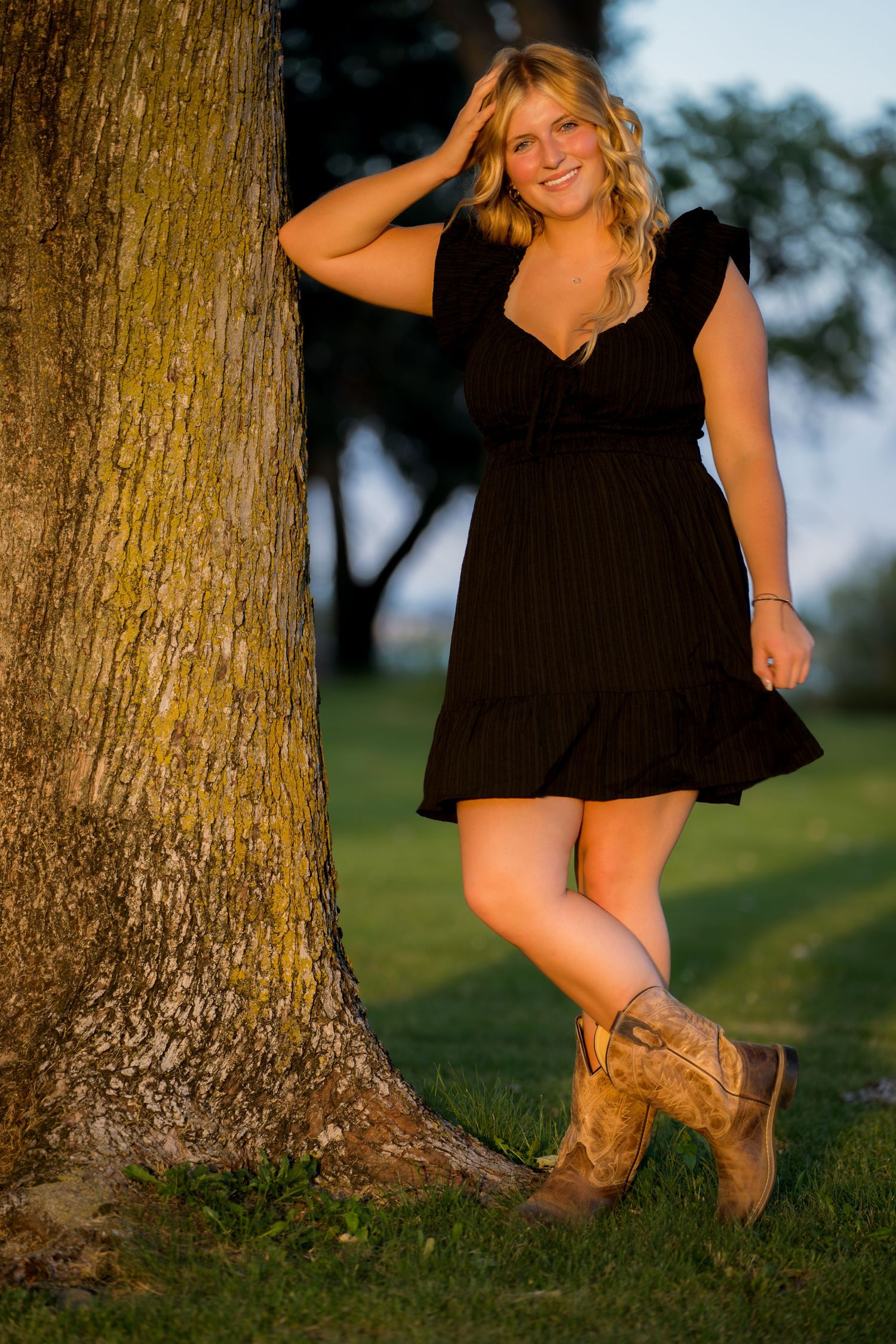 senior girl in dress poses by tree wearing cowboy boots.