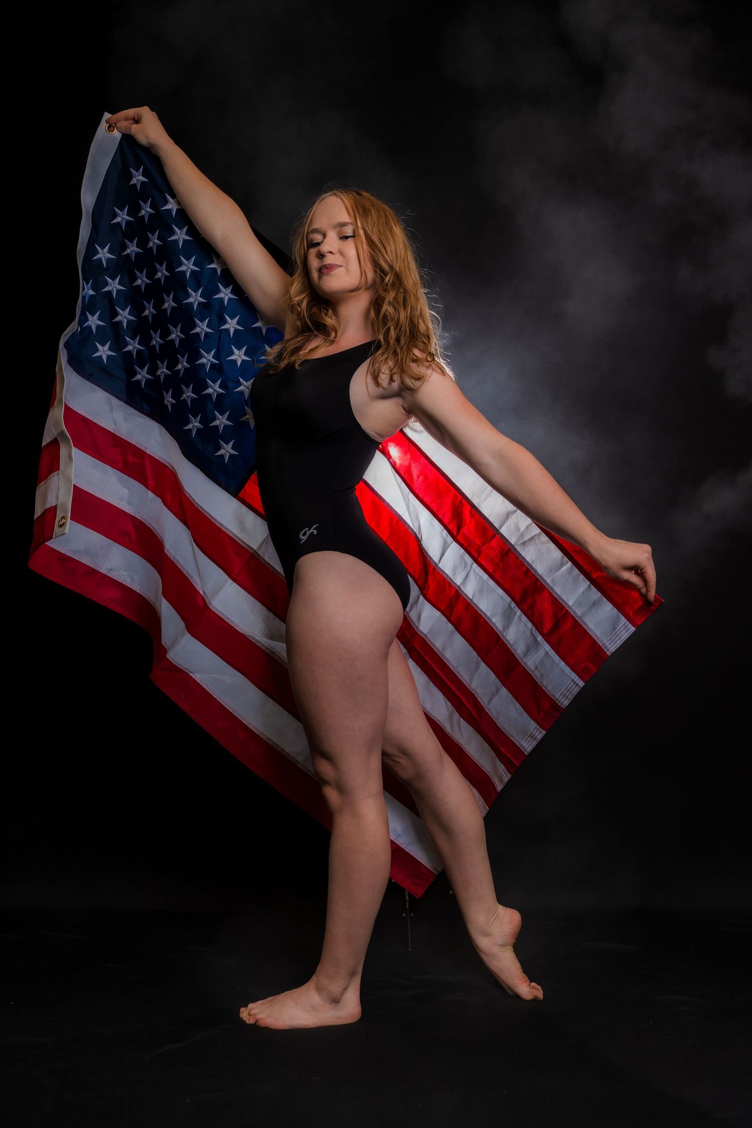 A gymnast in a black leotard poses gracefully with an American flag against a dark background.