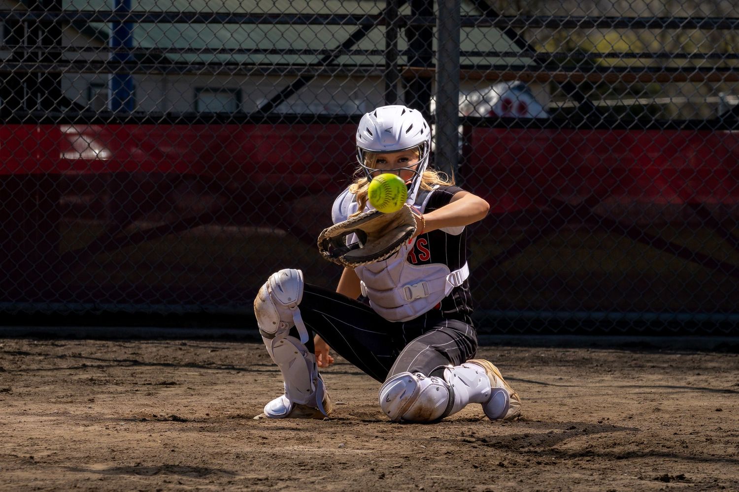 Softball catcher in full gear crouching to catch a yellow ball during a game on a dirt field.