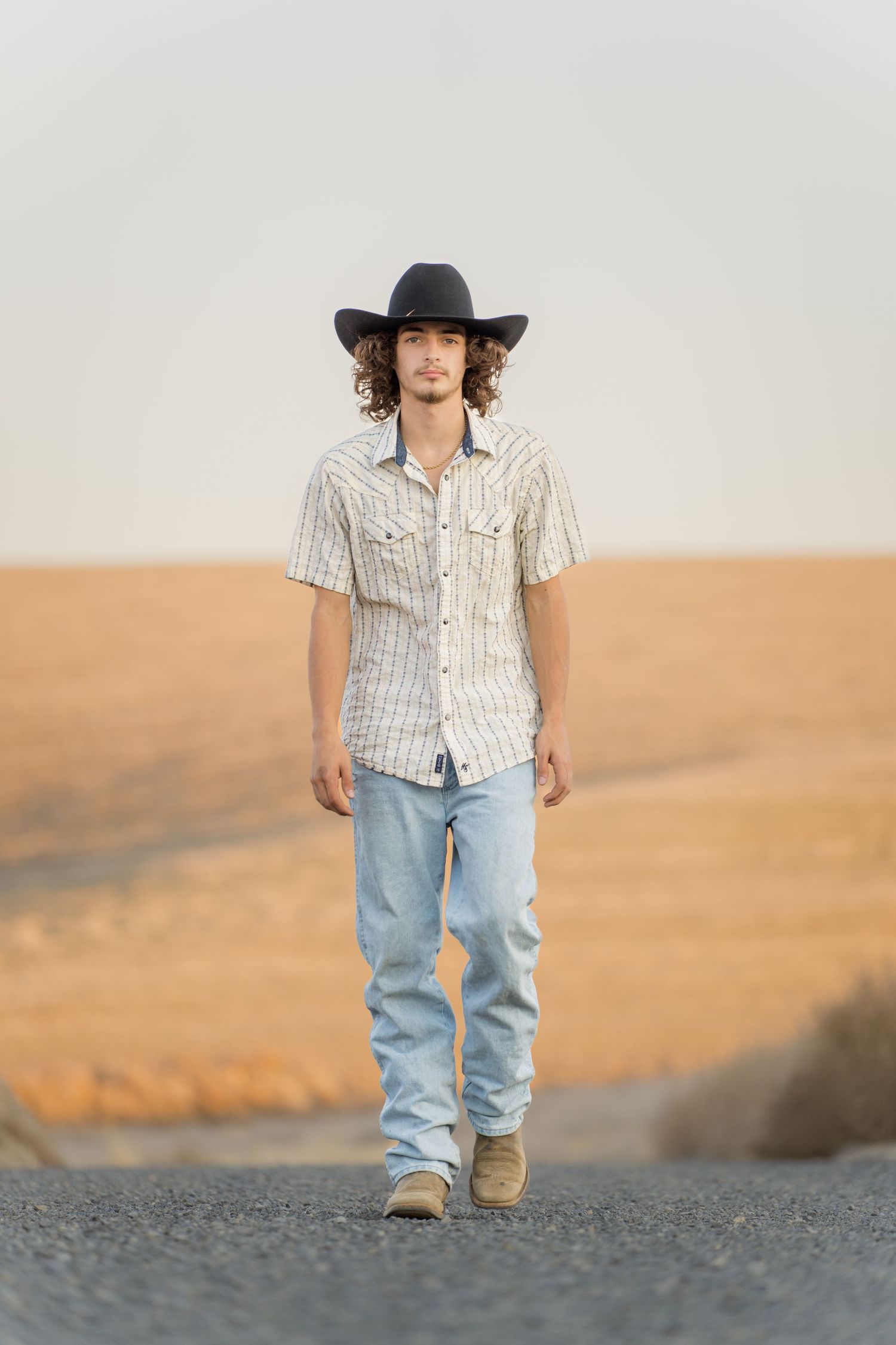 A young man in a cowboy hat, striped shirt and jeans stands on a dirt road at sunset.