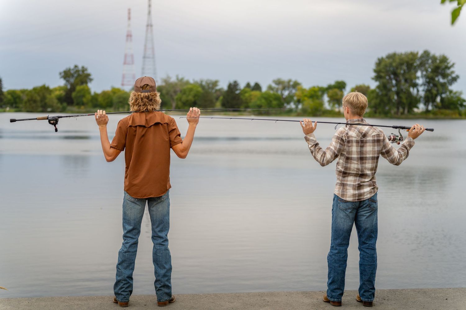 Two brothers stand at the edge of a lake holding fishing poles against a cloudy sky.