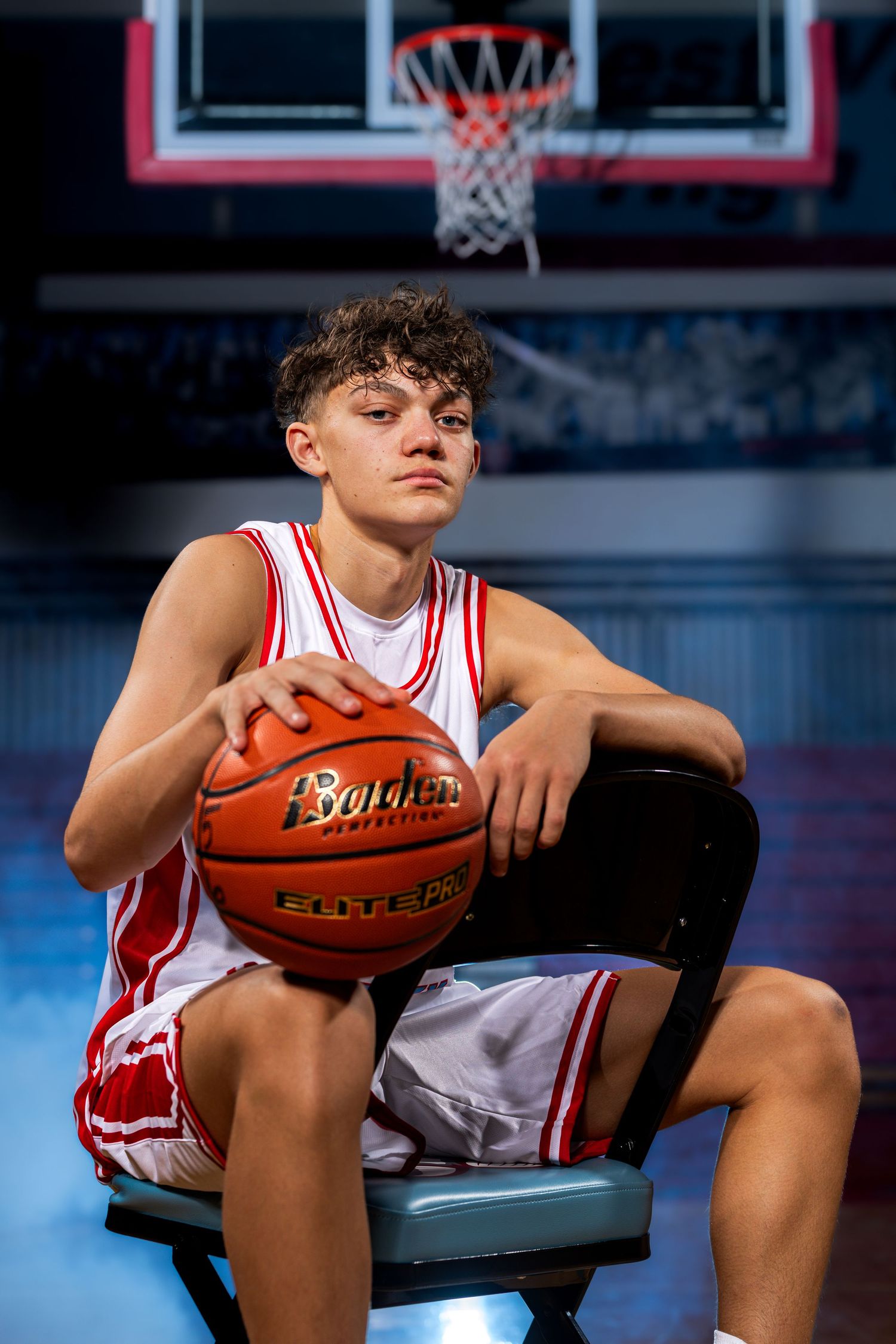 Young male basketball player in red and white uniform holding a Spalding ball, seated in front of a hoop.