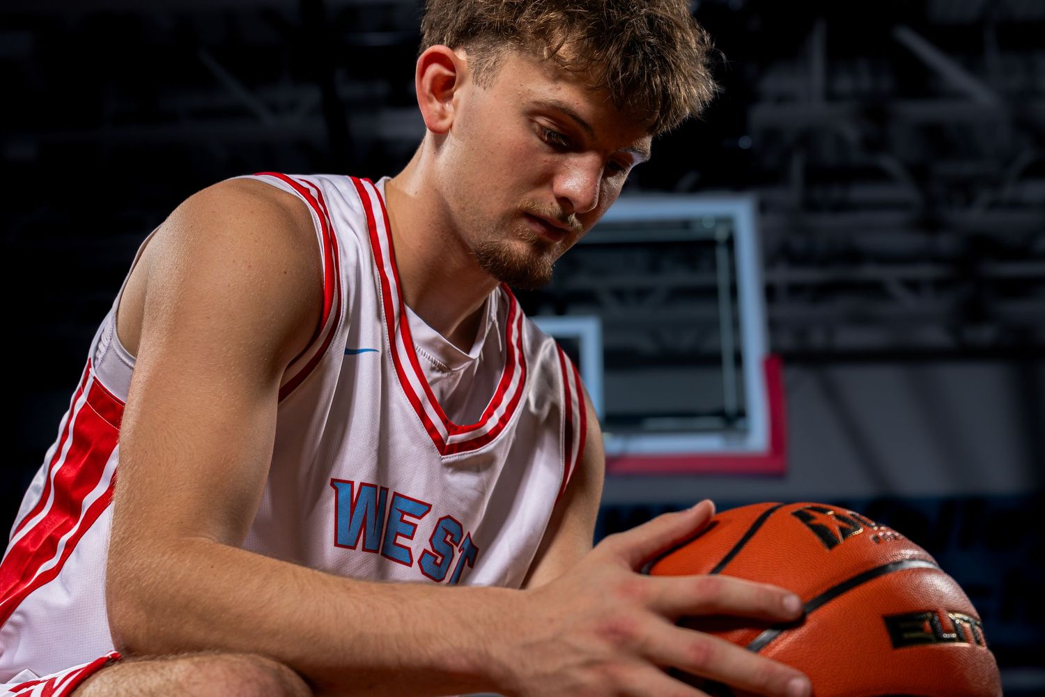 Basketball player in red and white jersey holds ball while looking down during dramatic indoor court photoshoot.