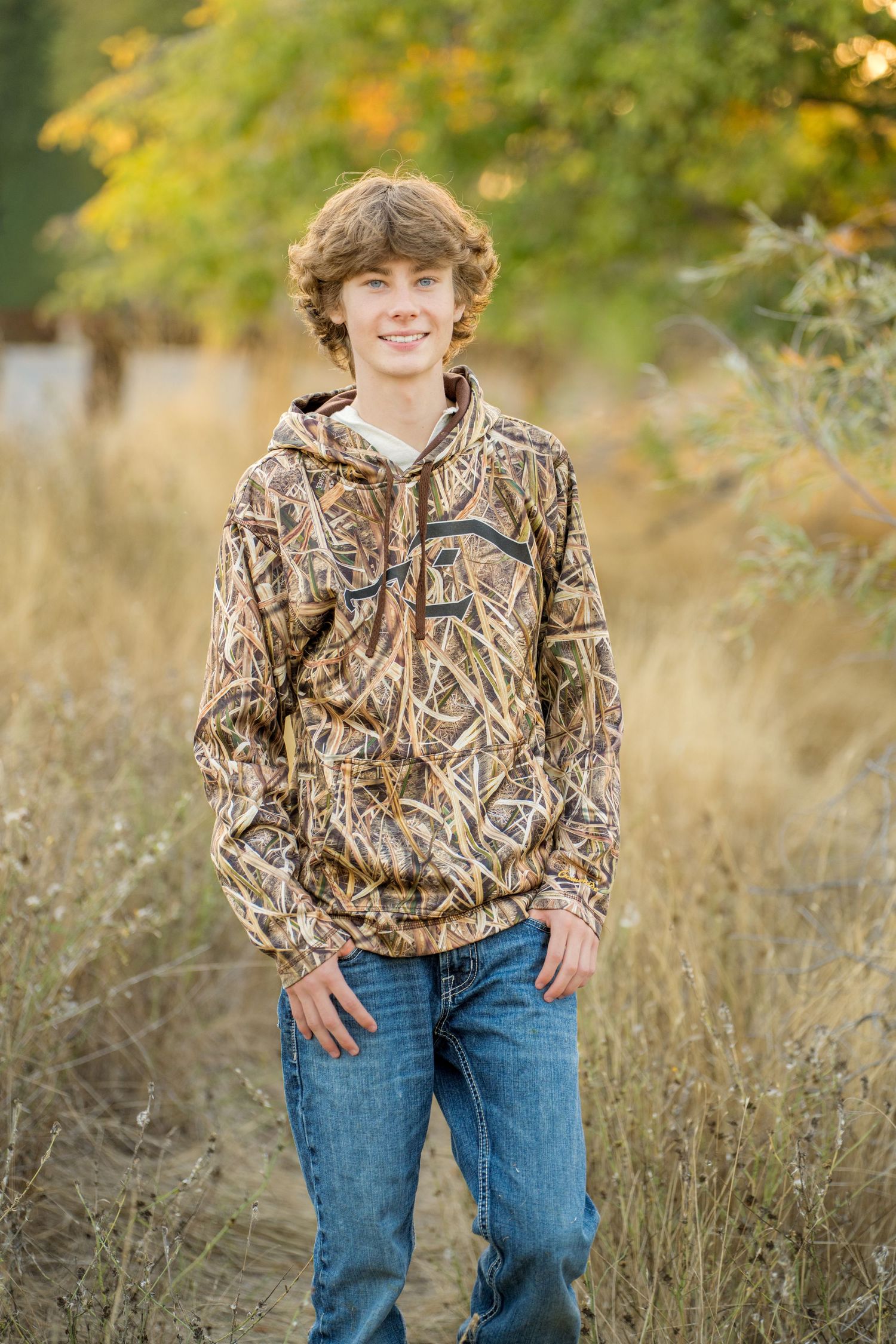 A young person in camouflage hunting attire poses outdoors in an autumn field setting.