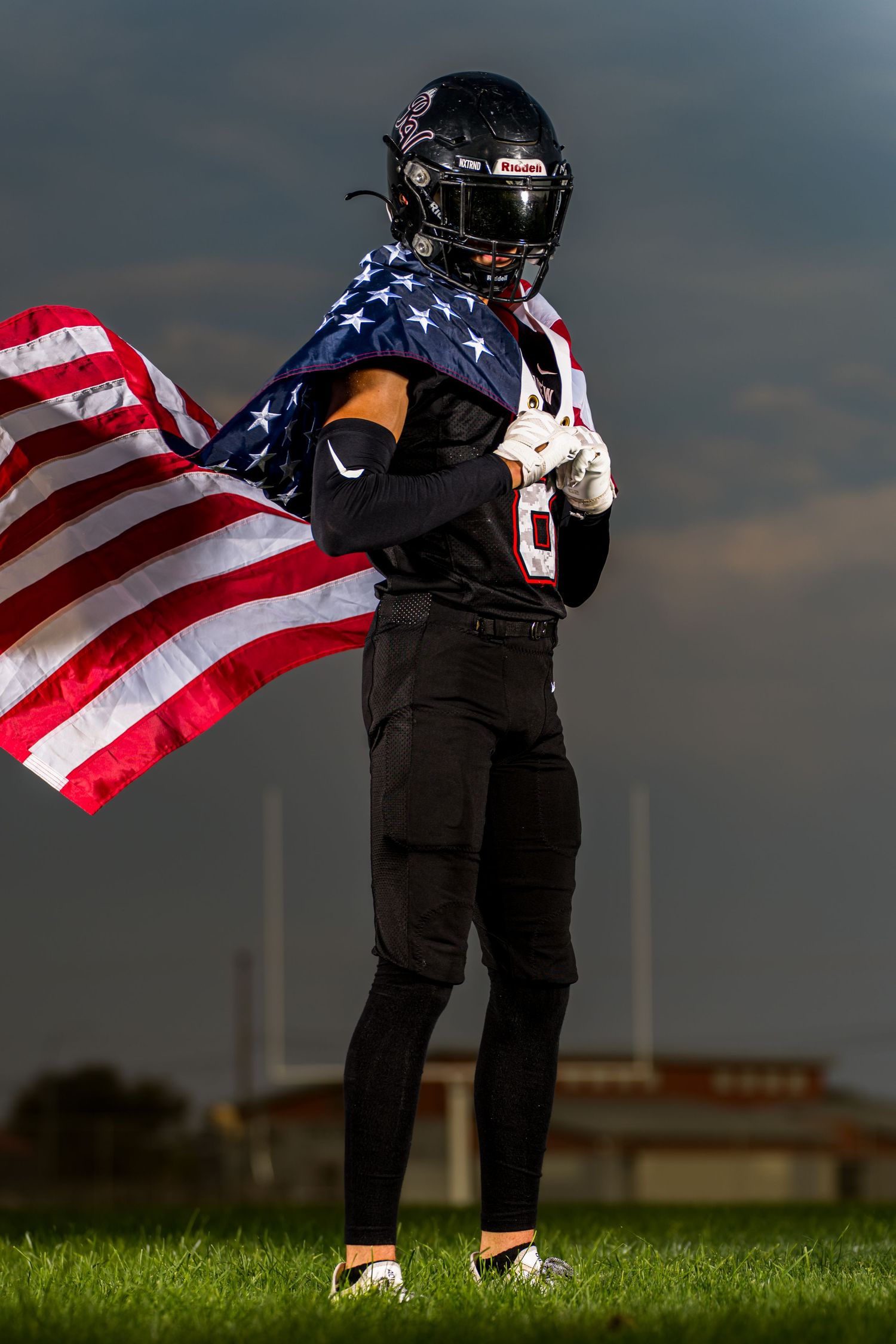 Football player in black uniform holds American flag against dramatic sky during sunset.