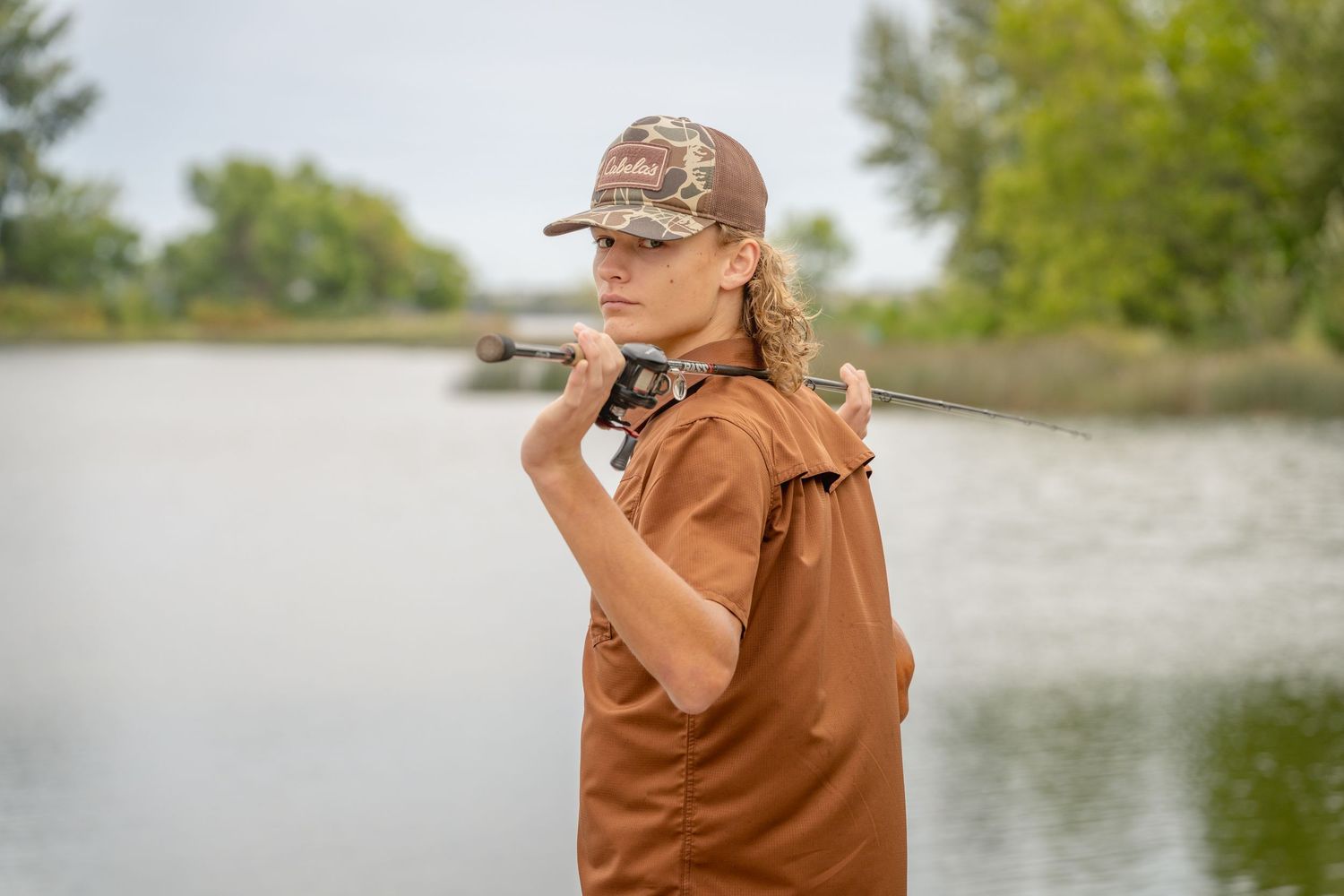 A senior boy in a brown jacket holding a fishing pole while posing near a lake.