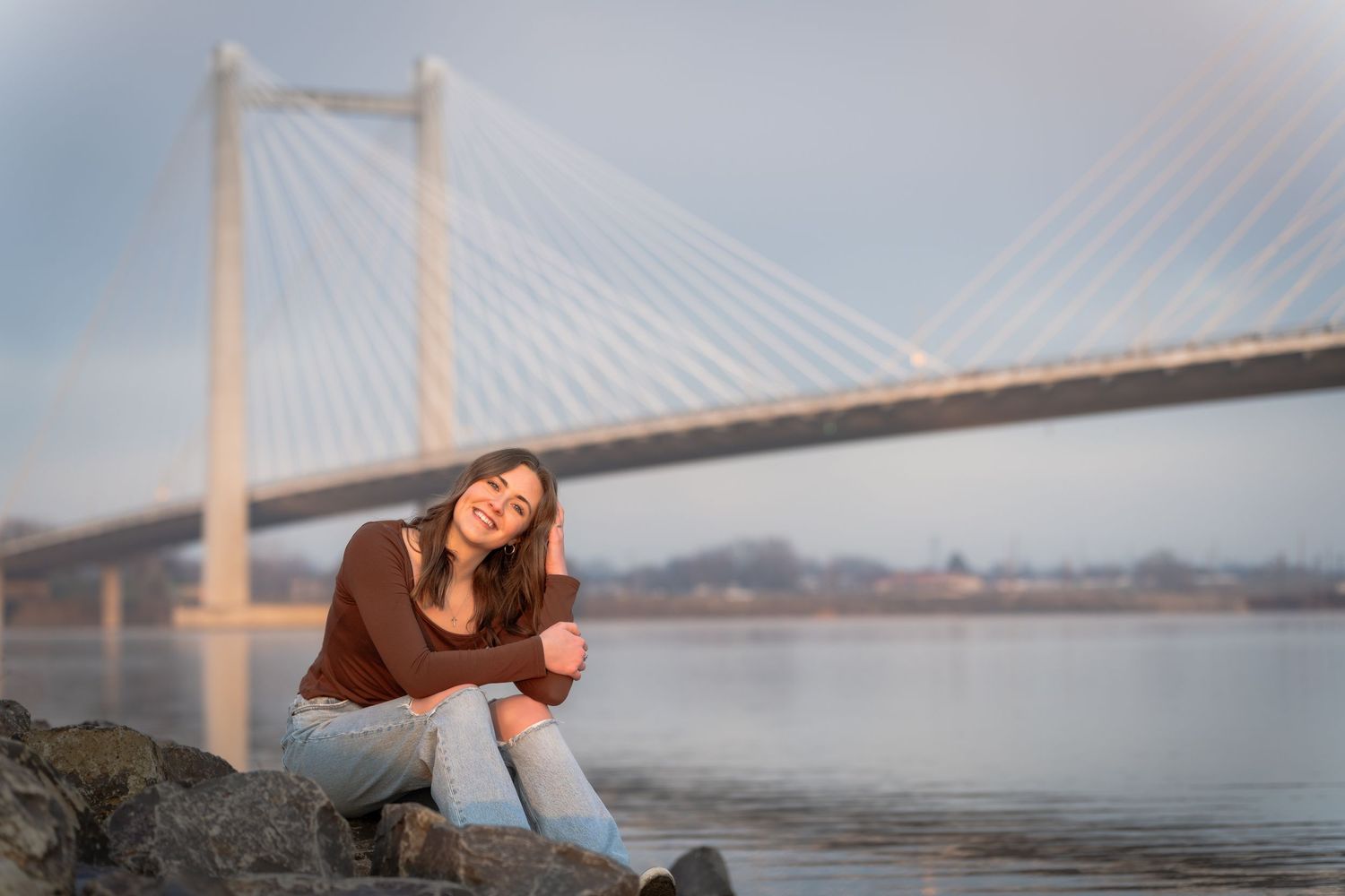 Young woman sitting on rocks by a river smiling, with a cable-stayed bridge in the misty background.