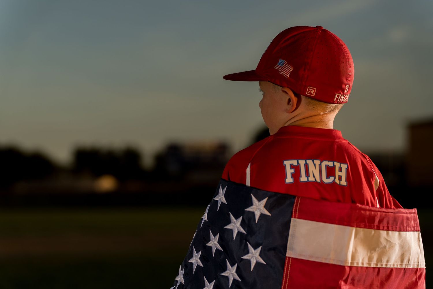 Youth baseball player wearing American flag over jersey stands against sunset sky.